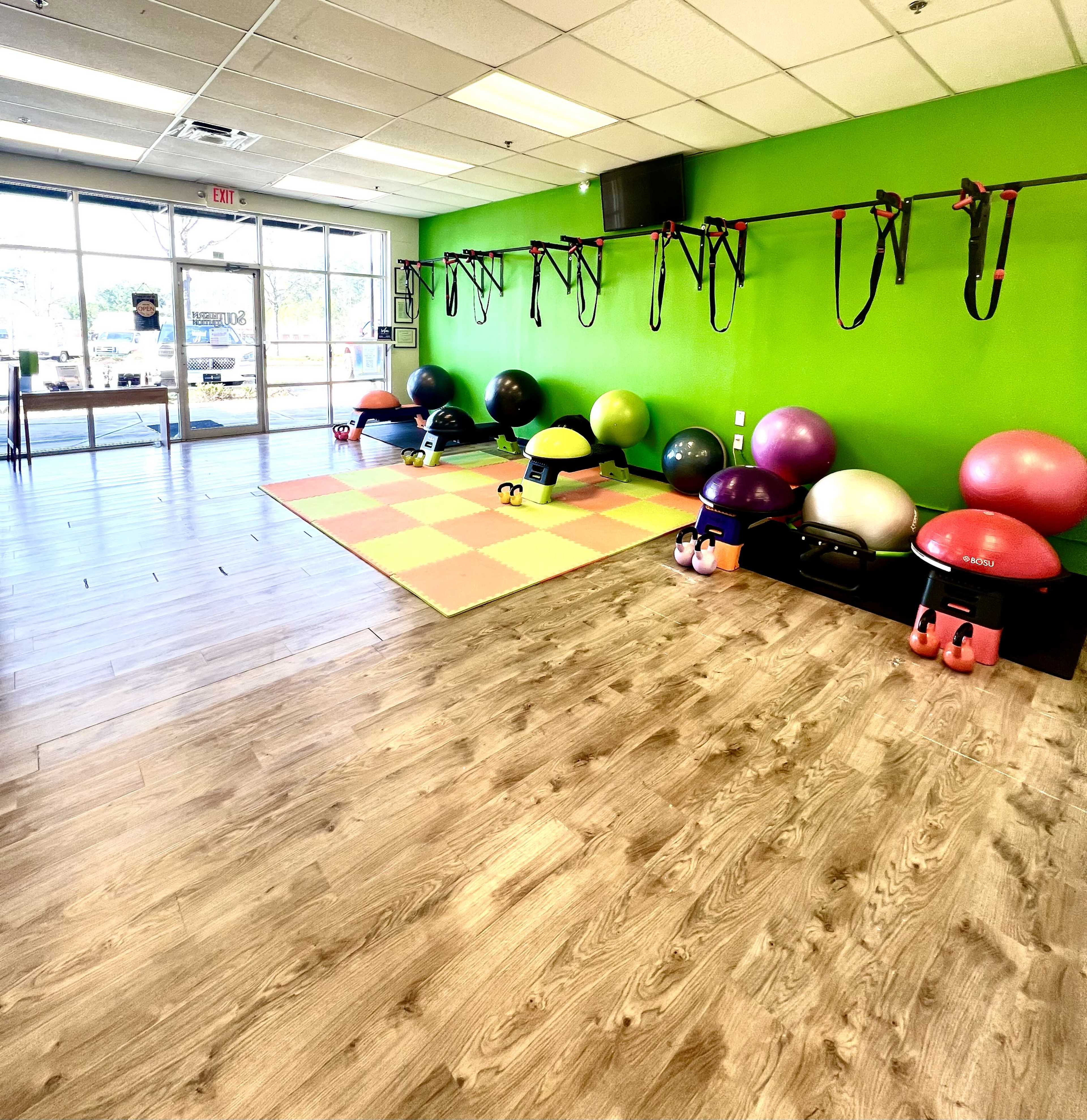 A fitness studio with various colored exercise balls and kettlebells arranged on a wooden floor, against a bright green wall.
