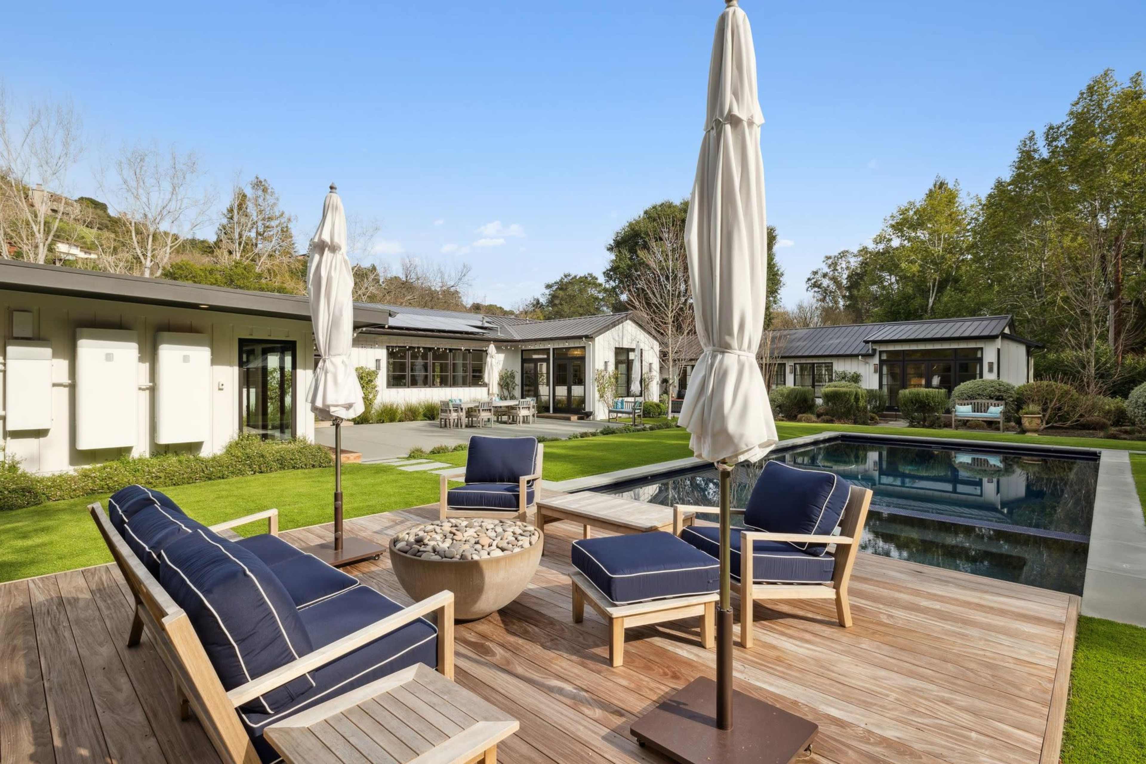 A modern patio features wooden furniture arranged around a circular fire pit, adjacent to a swimming pool surrounded by greenery and contemporary buildings.
