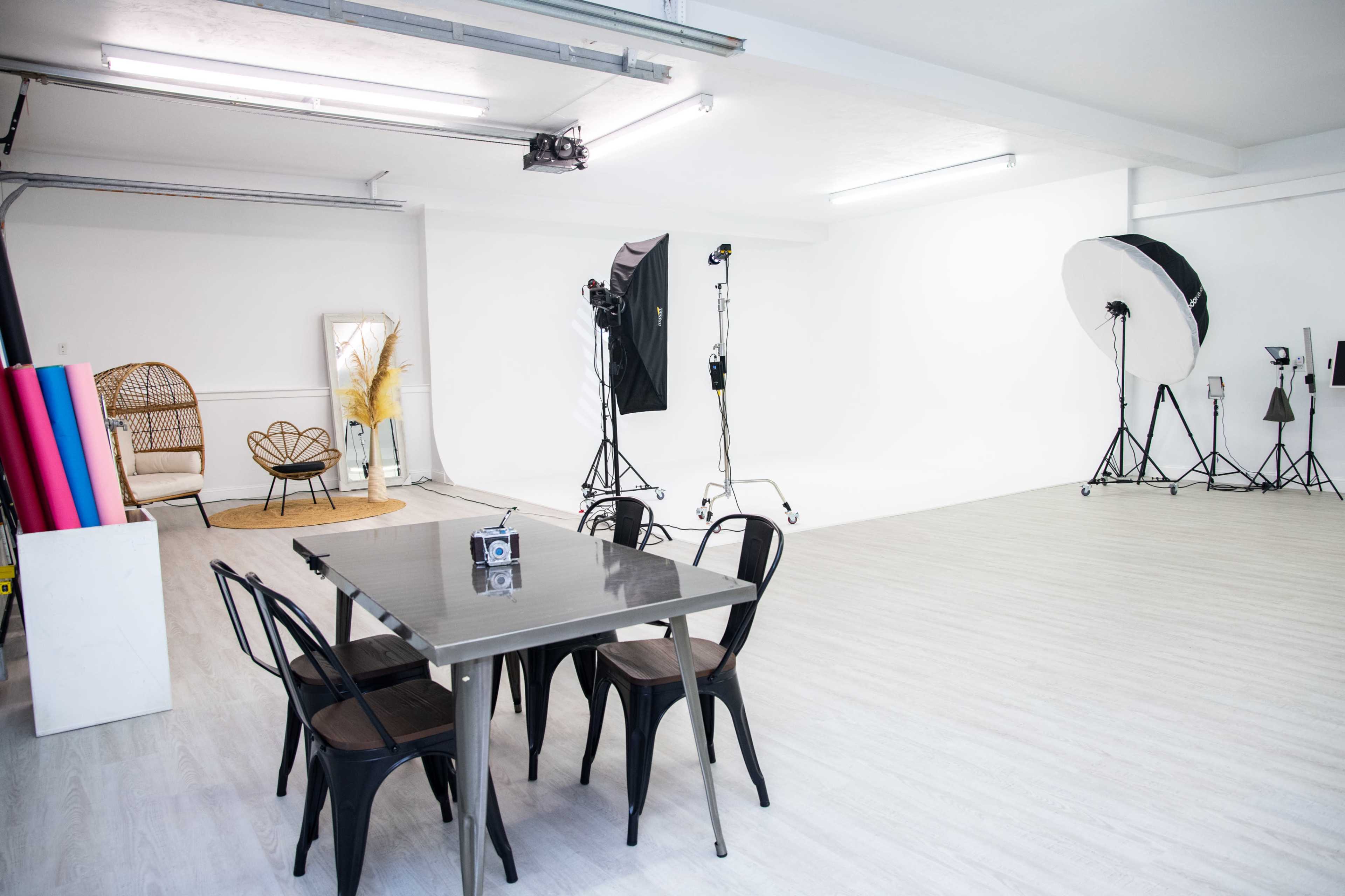 The image shows a photography studio with a large white backdrop, a table with black chairs, and various lighting equipment positioned around the space.