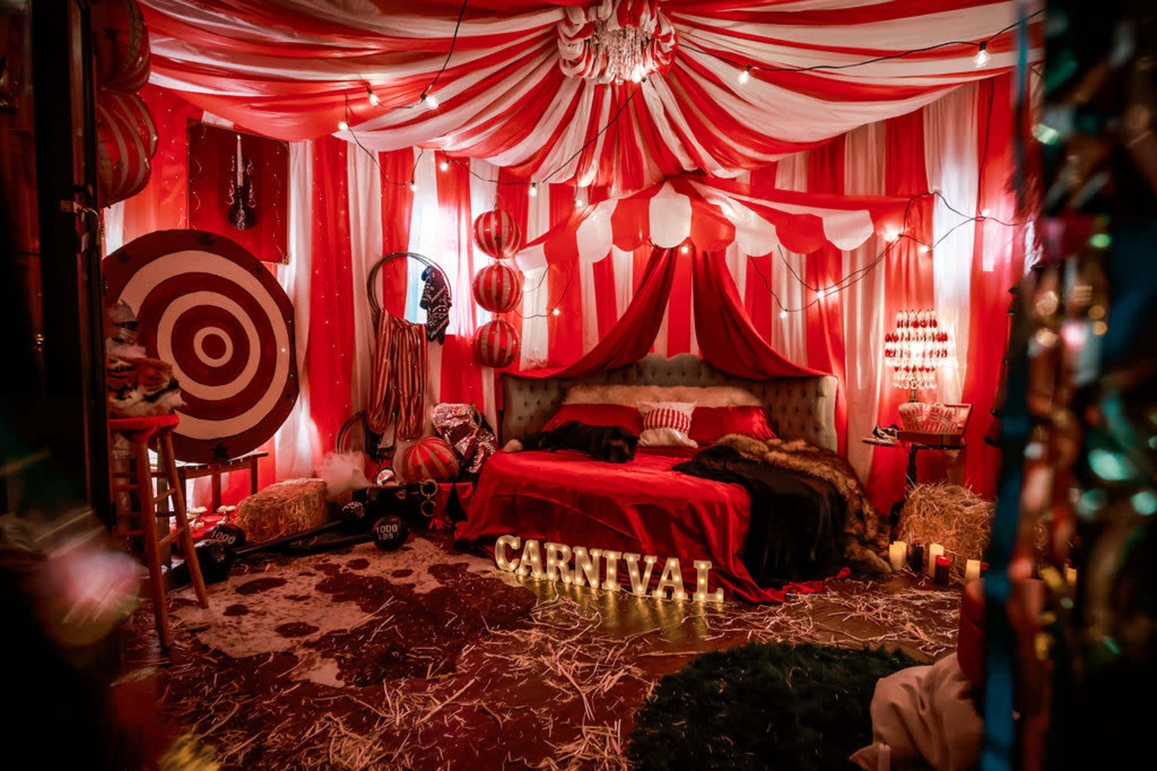 The image shows a vibrant carnival-themed bedroom decorated with red and white striped drapes, a bed with matching linens, and various carnival props like targets and lights, all set on a straw-covered floor.