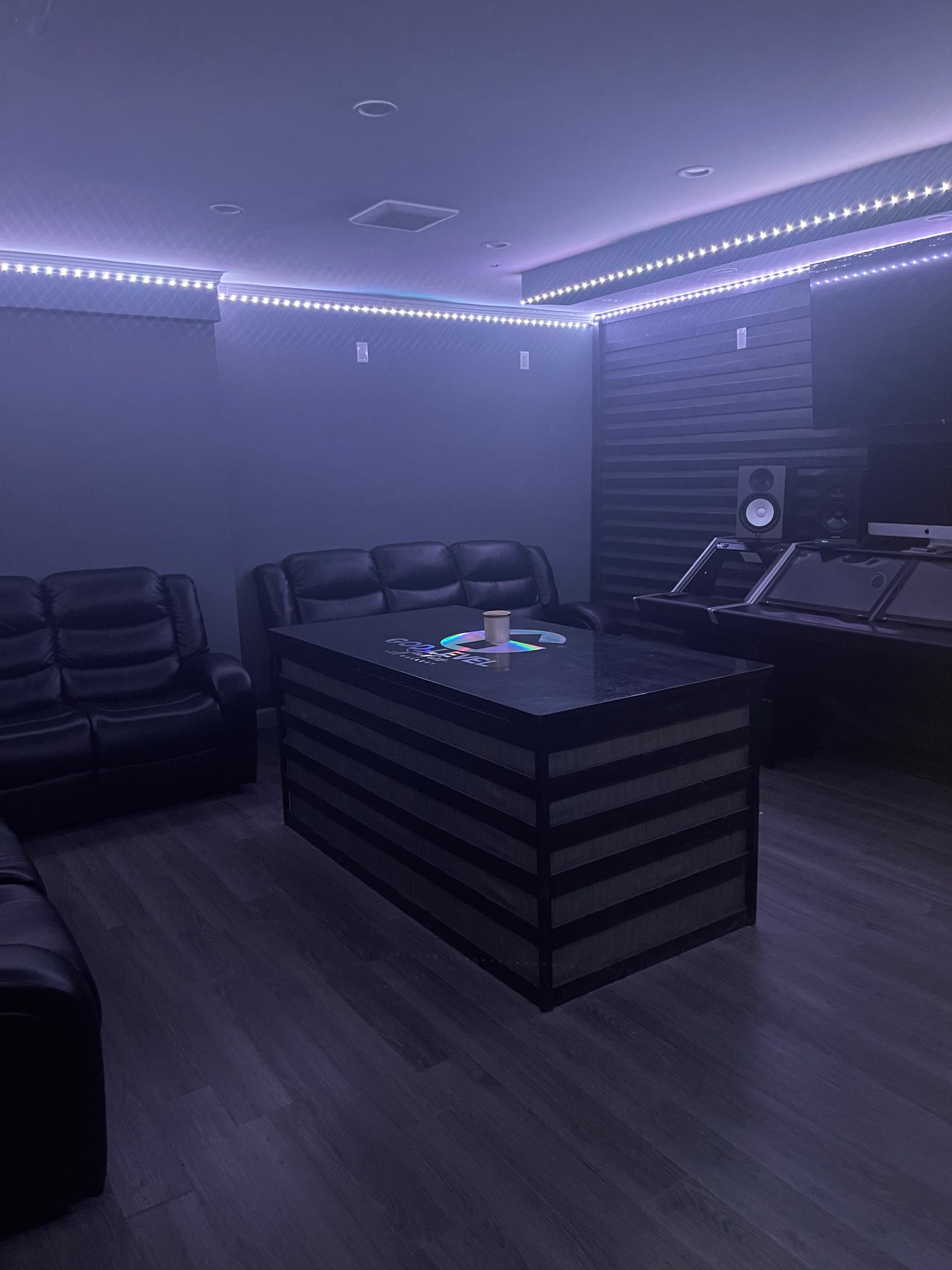 A dimly lit lounge area with black leather sofas, a central table, and equipment for audio production.
