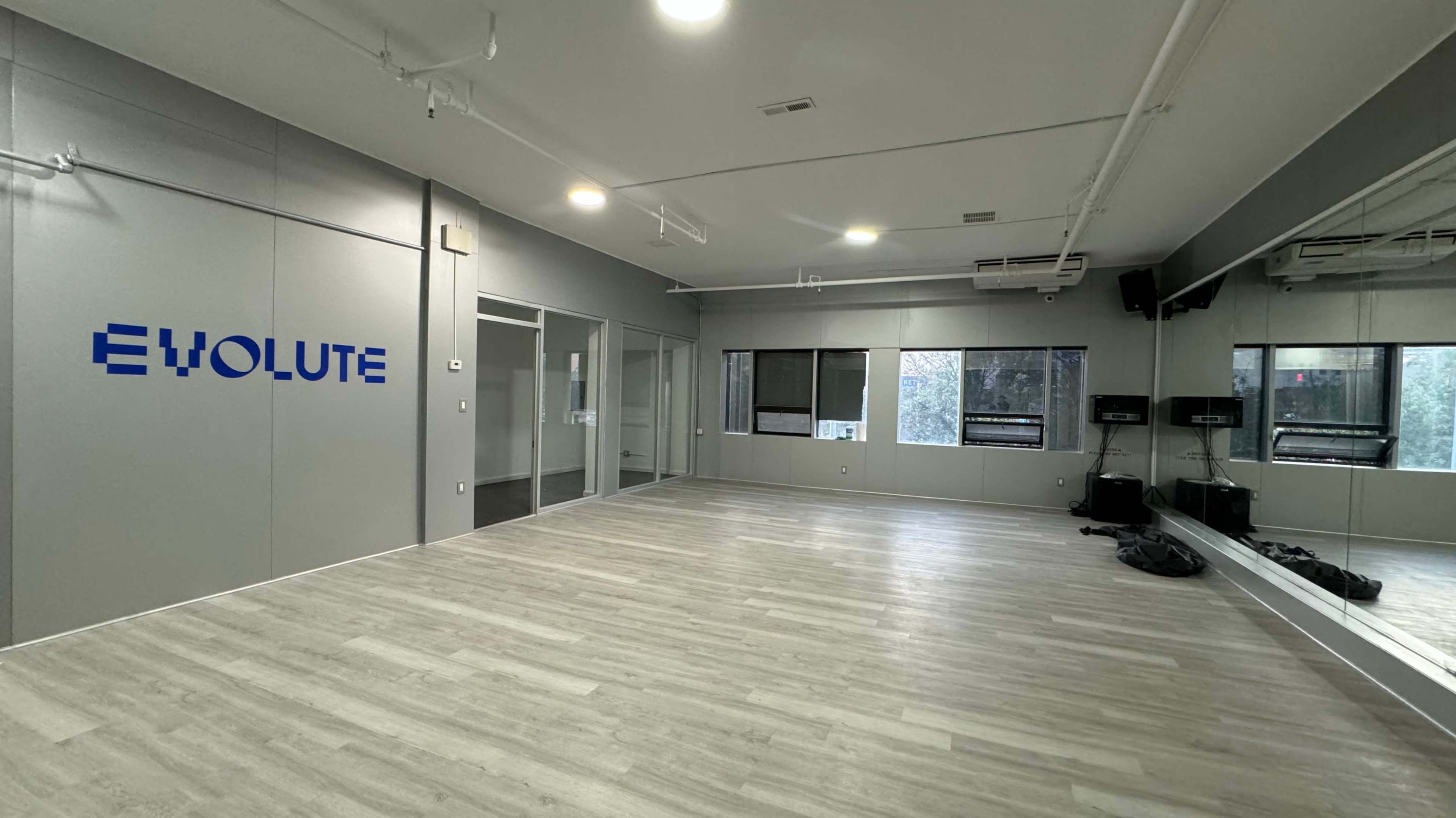 The image shows a spacious, empty room with light wooden flooring and a gray wall featuring the word "EVOLUTE" in blue, along with glass partitions and large windows.