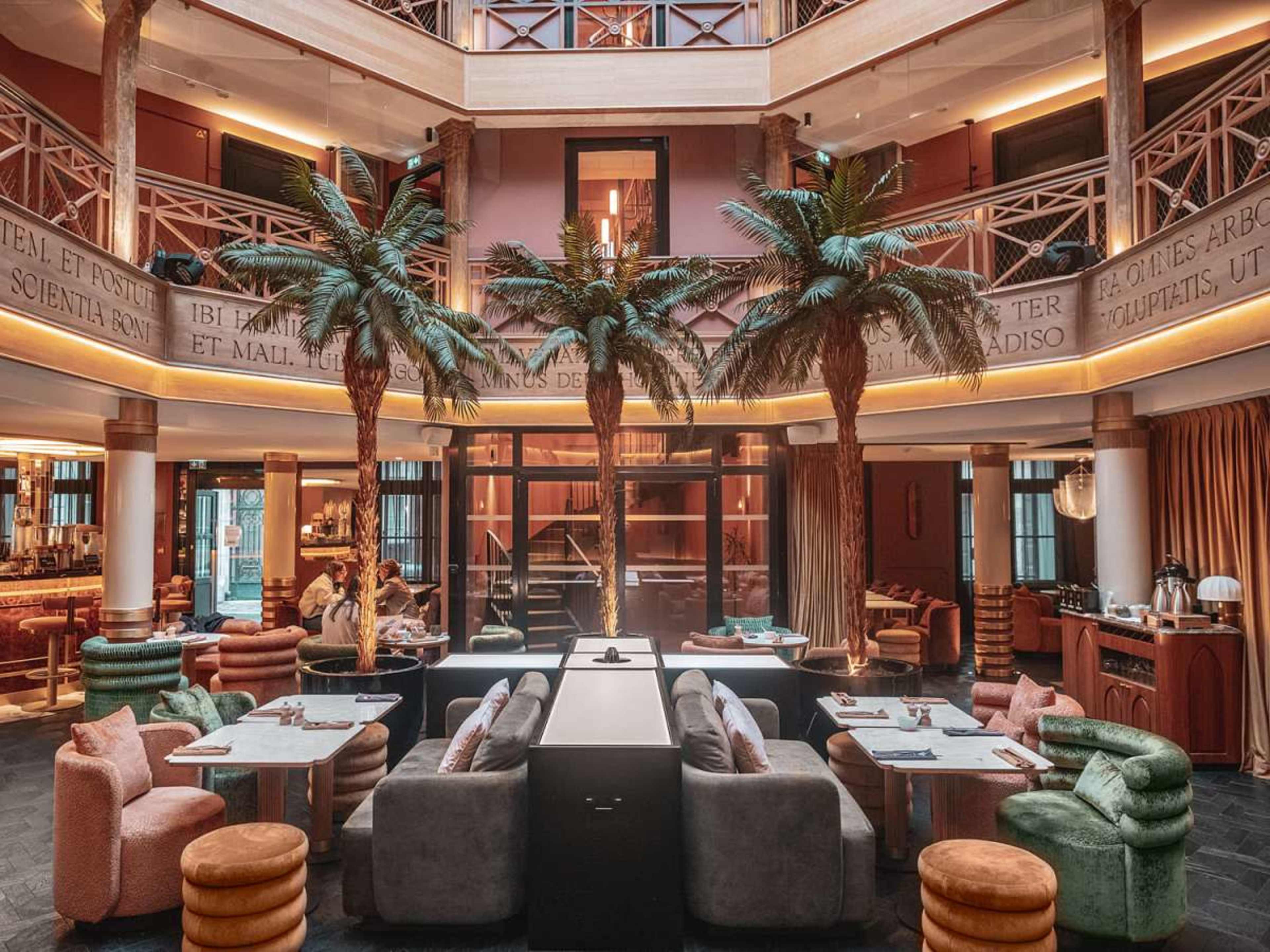 The image shows a modern interior of a space with palm trees, colorful seating arrangements, and a central bar area, creating an inviting atmosphere.