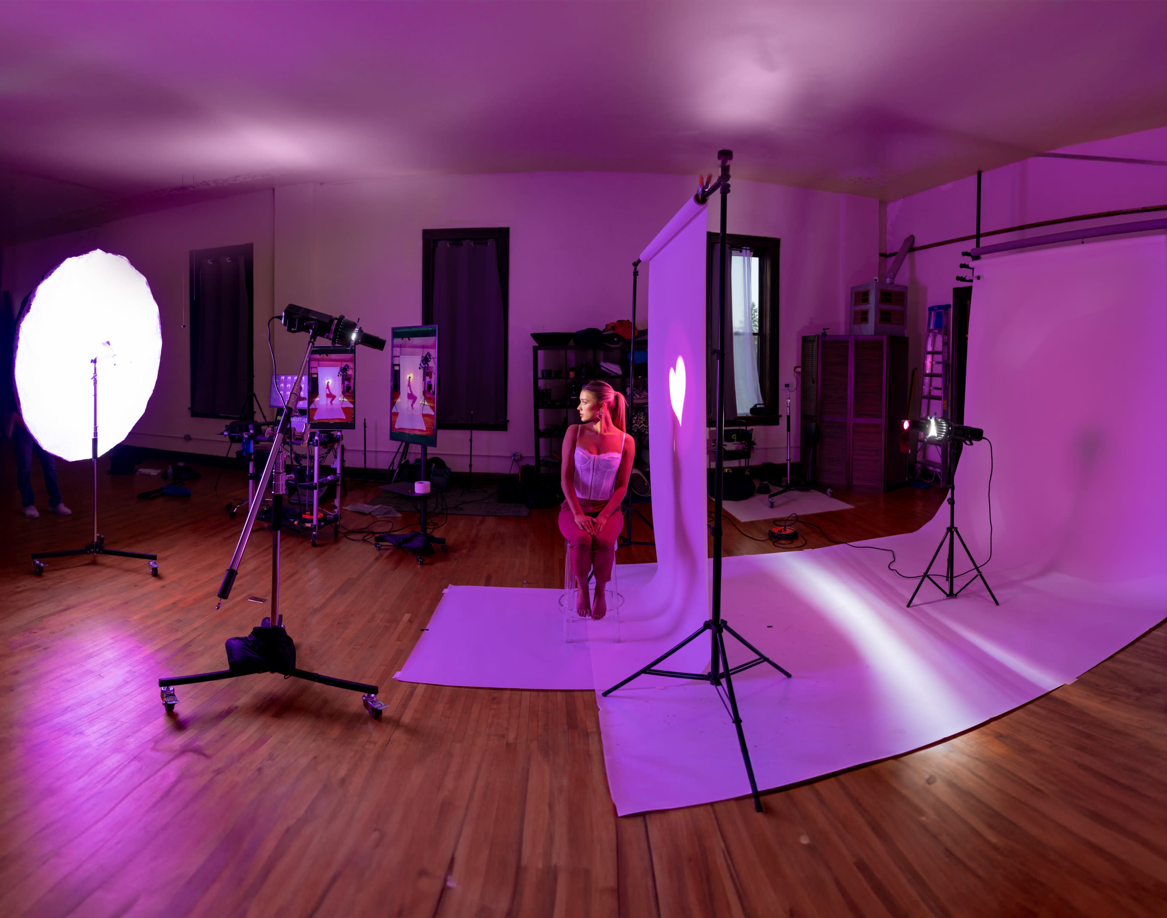 A photoshoot setup features a model sitting under bright lights, surrounded by backdrops and photography equipment in a studio with wooden flooring.