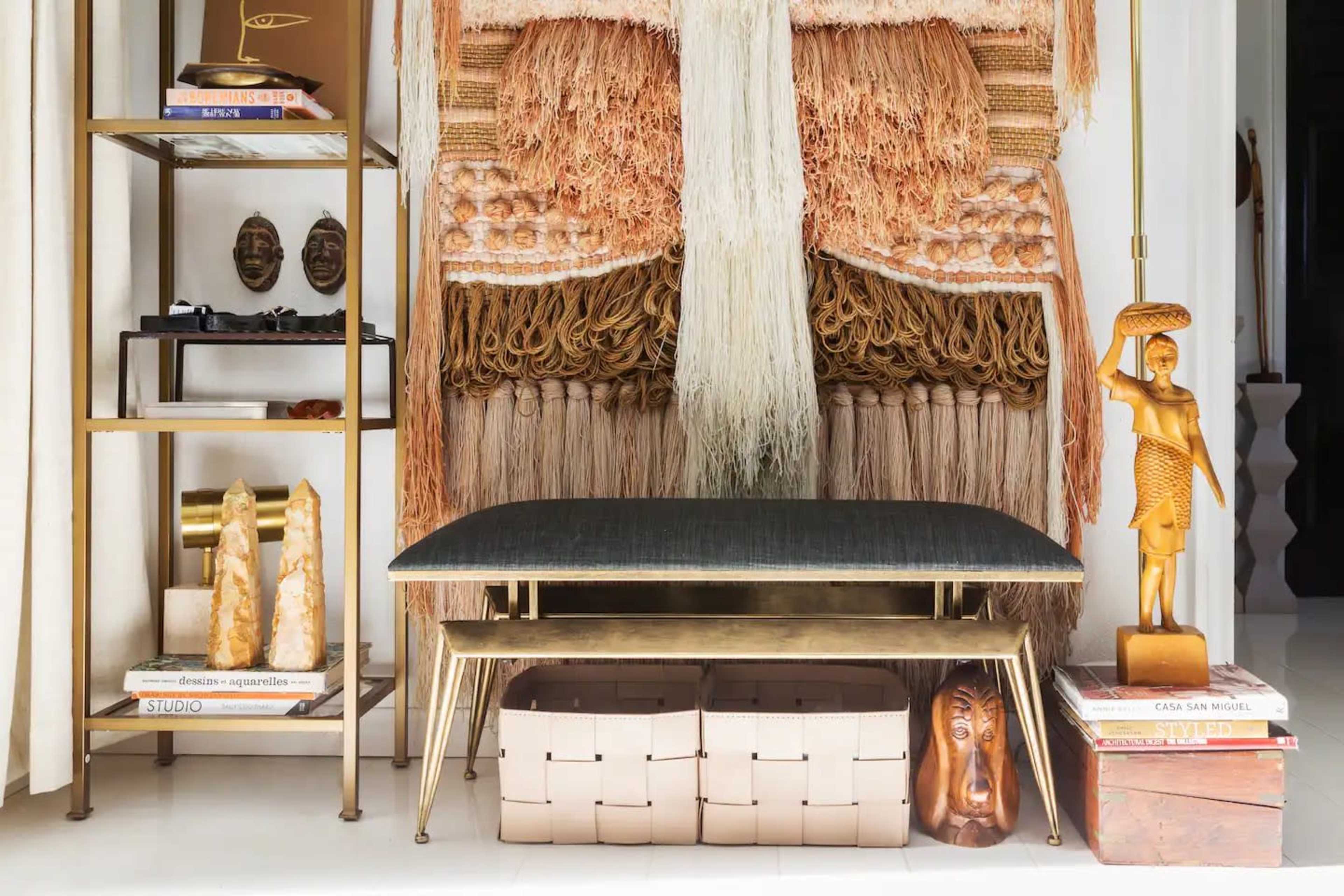 A decorative interior scene with a fringed wall hanging behind a sleek, metallic coffee table, flanked by a shelf displaying various sculptures and books.
