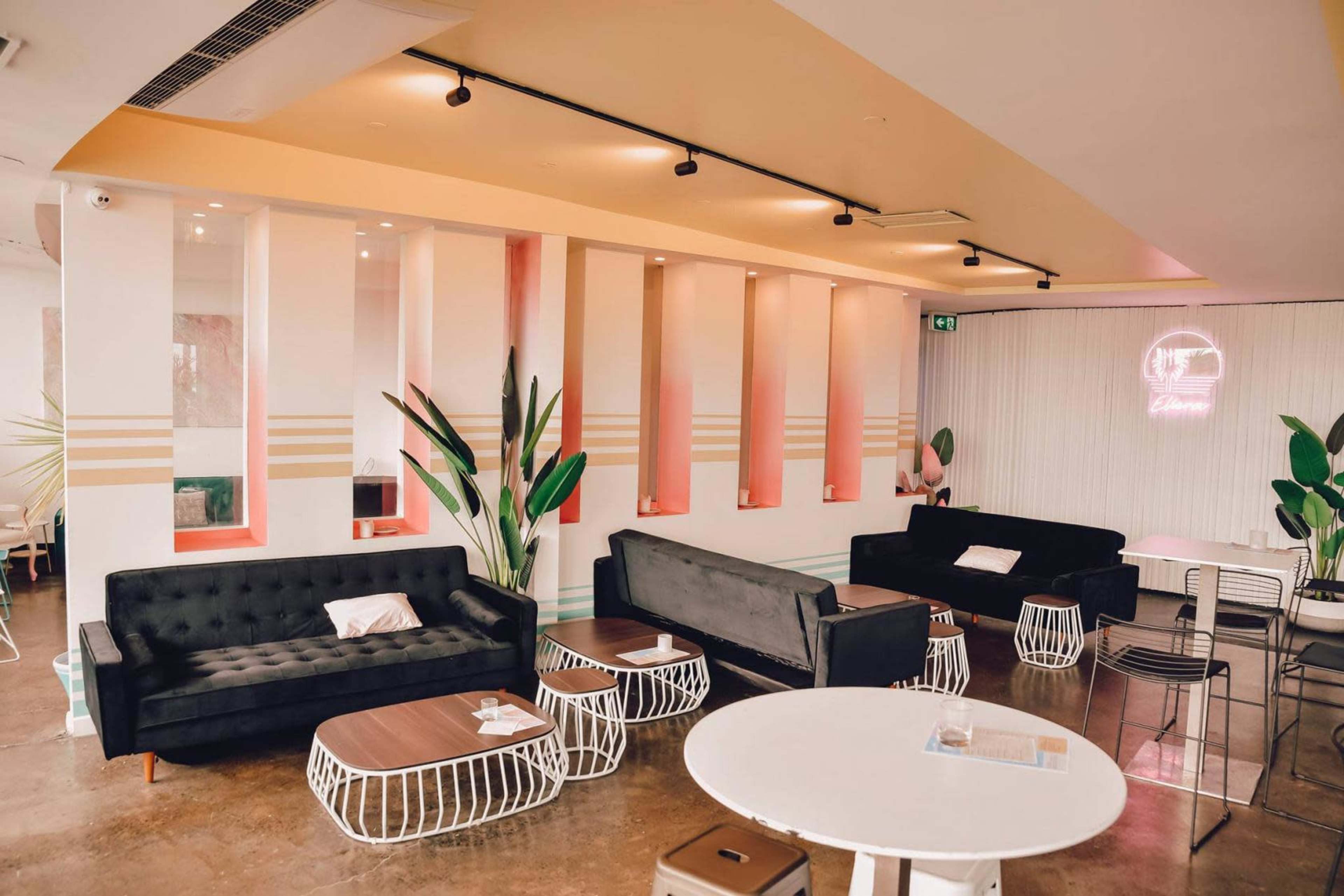 A modern café interior features black sofas, wooden tables, and decorative plants against a backdrop of pastel-colored walls.