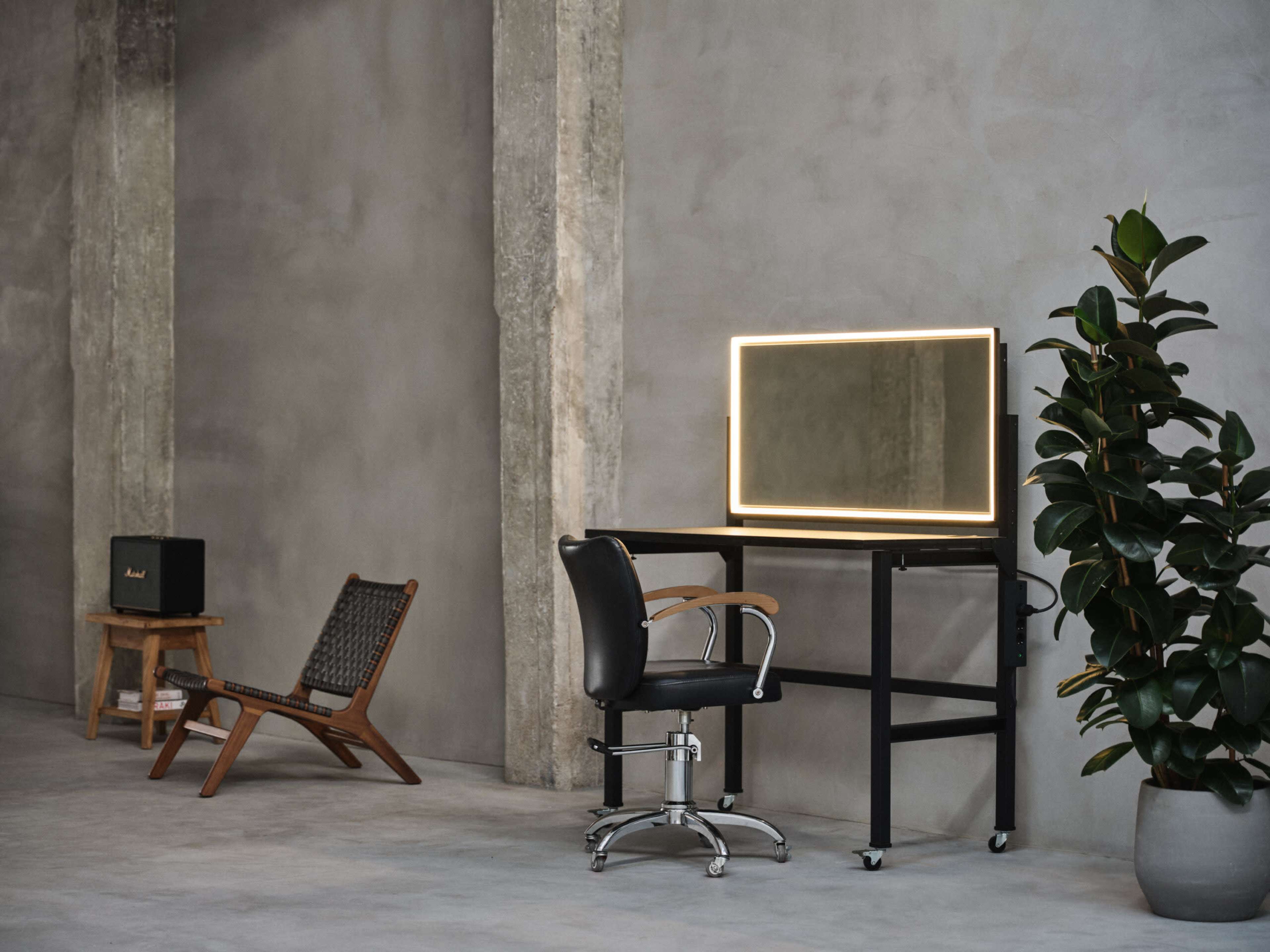 The image shows a minimalist interior with a desk featuring a mirror and a chair, a wooden lounge chair, a compact speaker, and a potted plant against a concrete wall.