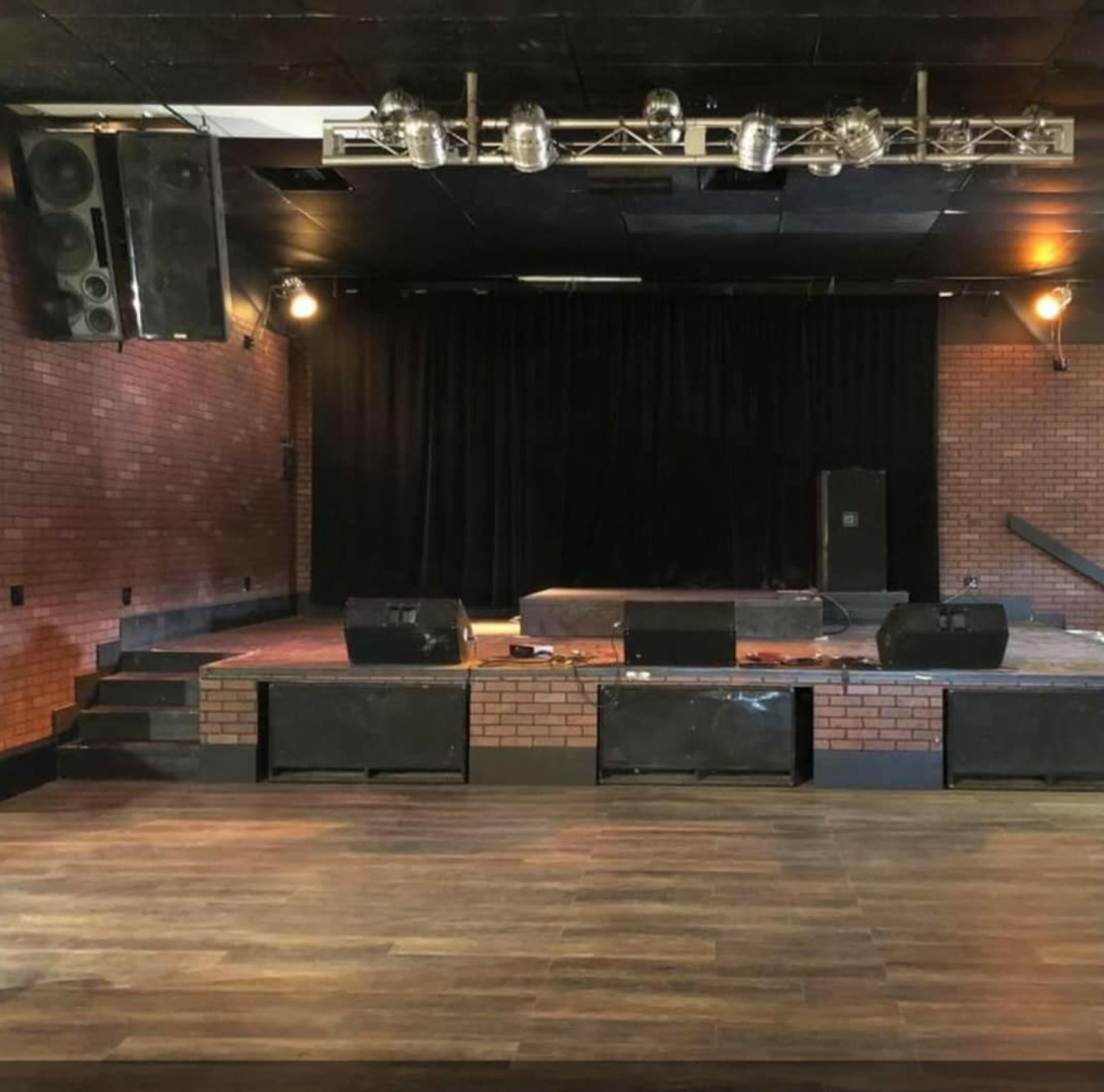 The image shows a stage area in a venue with a brick wall backdrop, wooden flooring, and sound equipment set up.