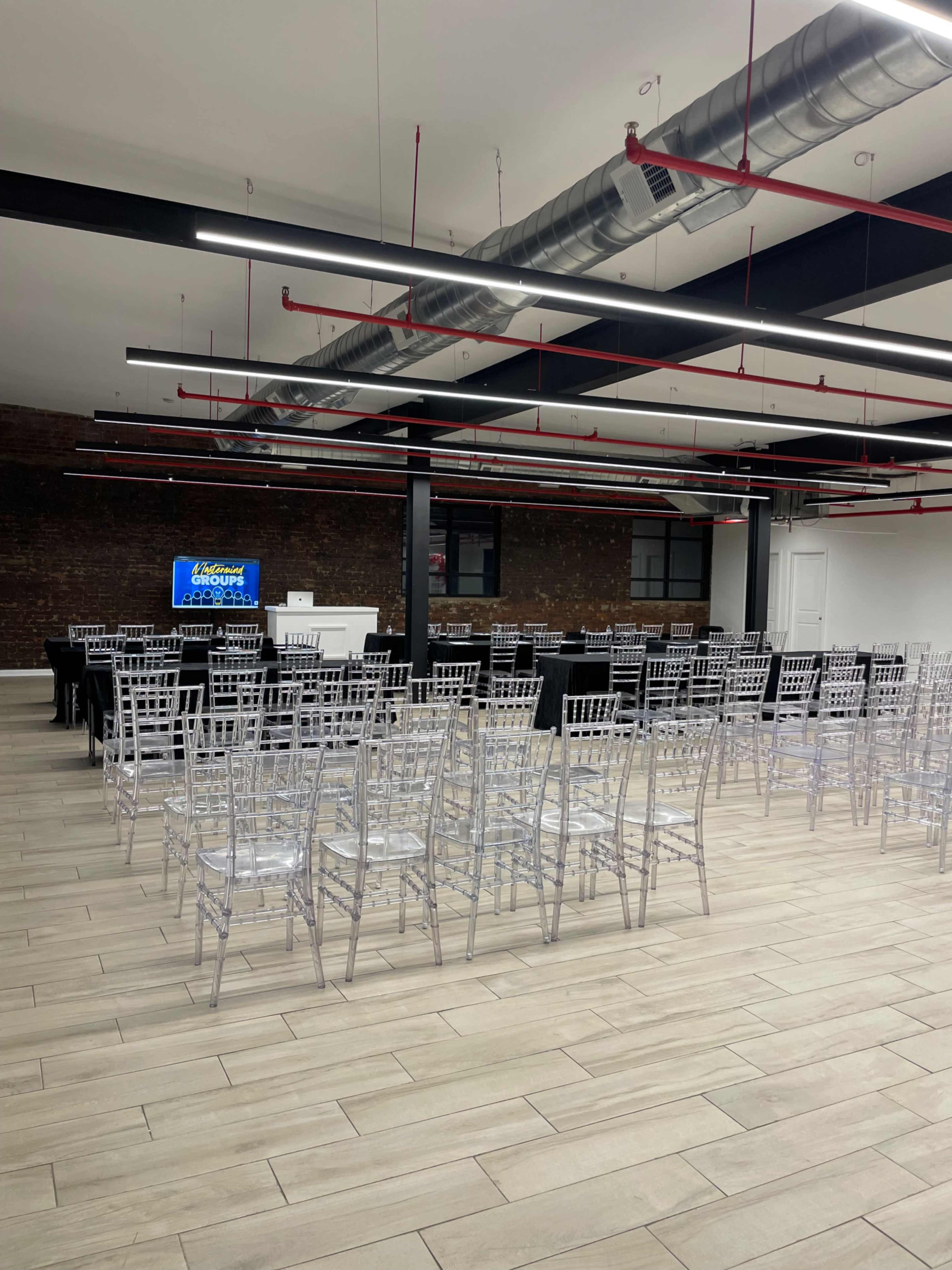 The image shows a spacious event room set up with multiple rows of clear chairs facing a presentation area.
