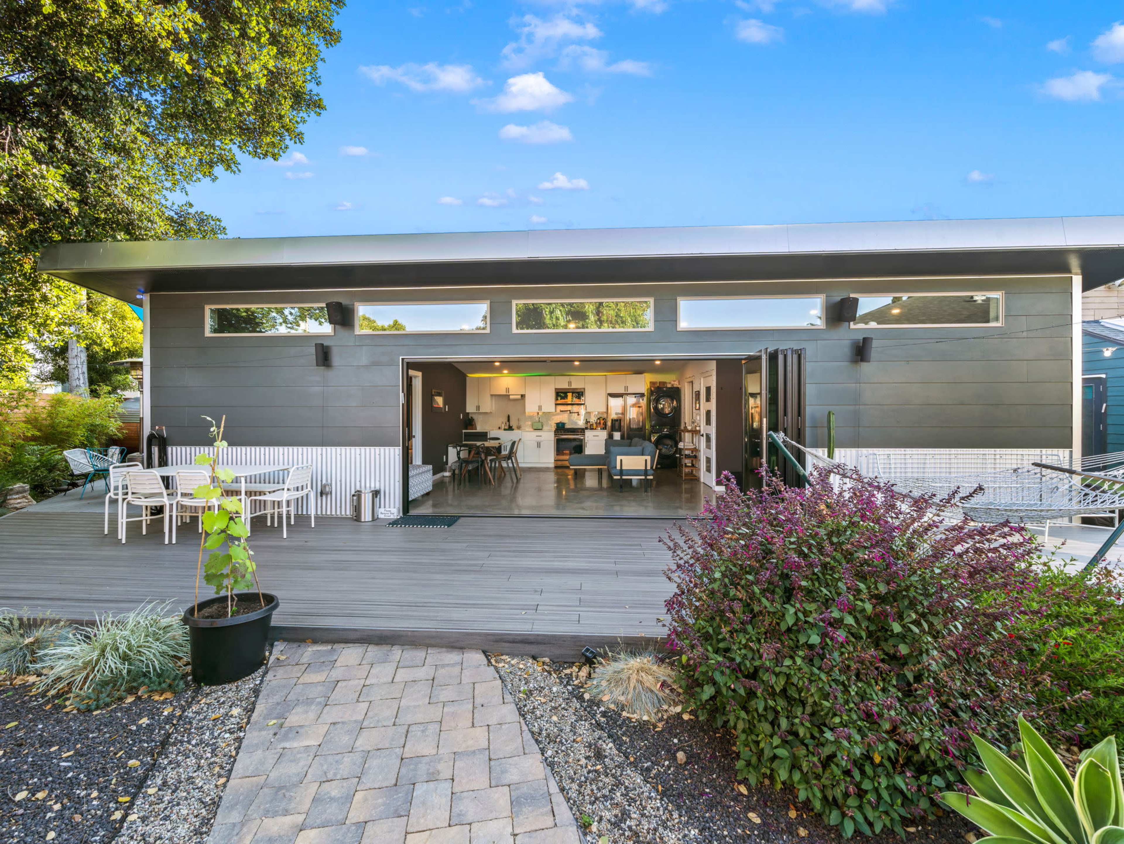 A modern house with large sliding glass doors opens to a wooden deck and garden, featuring minimalistic outdoor furniture and lush greenery.
