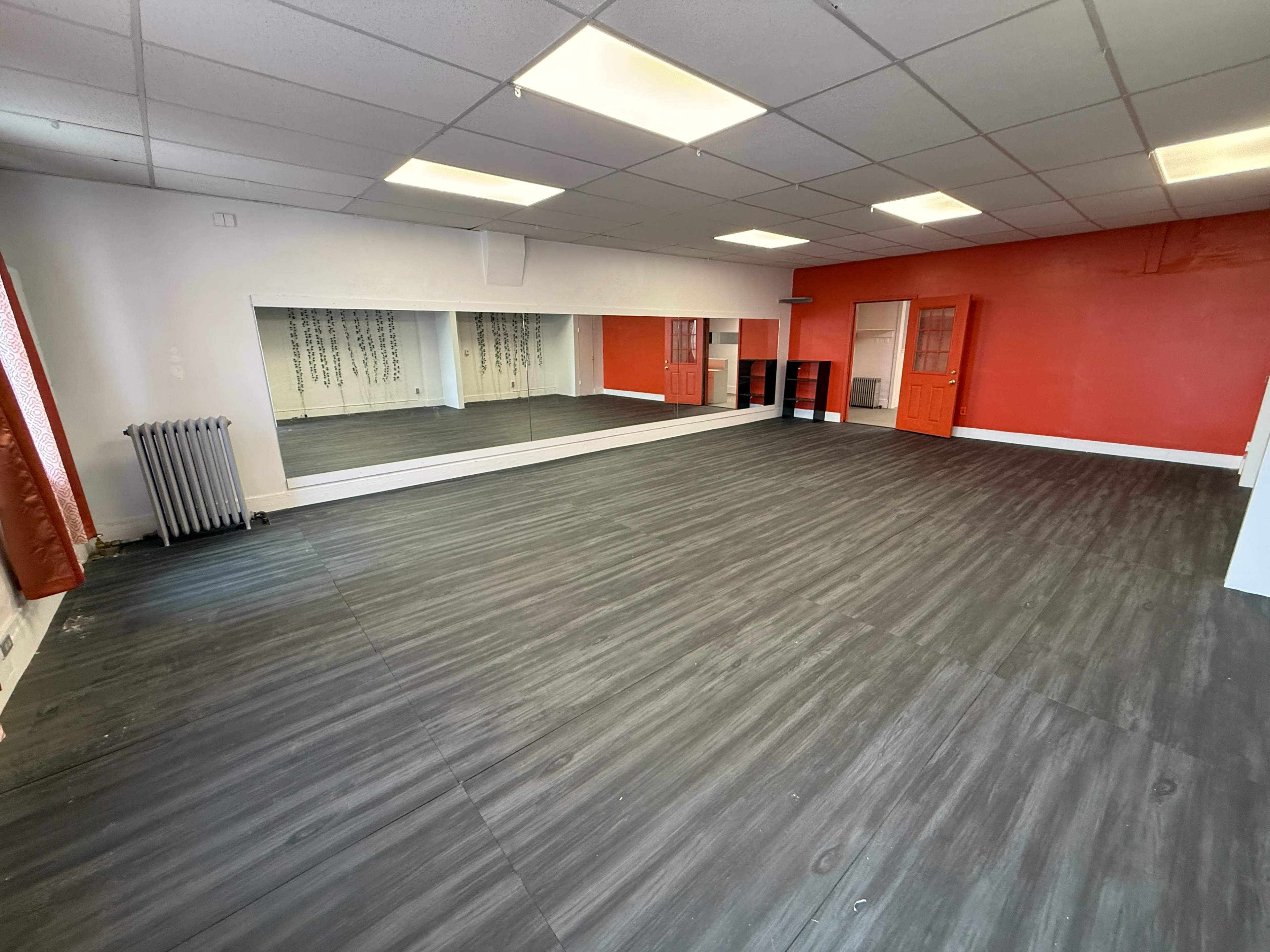 The image shows an empty room with a hardwood floor, red walls, a large mirror on one side, and a radiator in the corner.