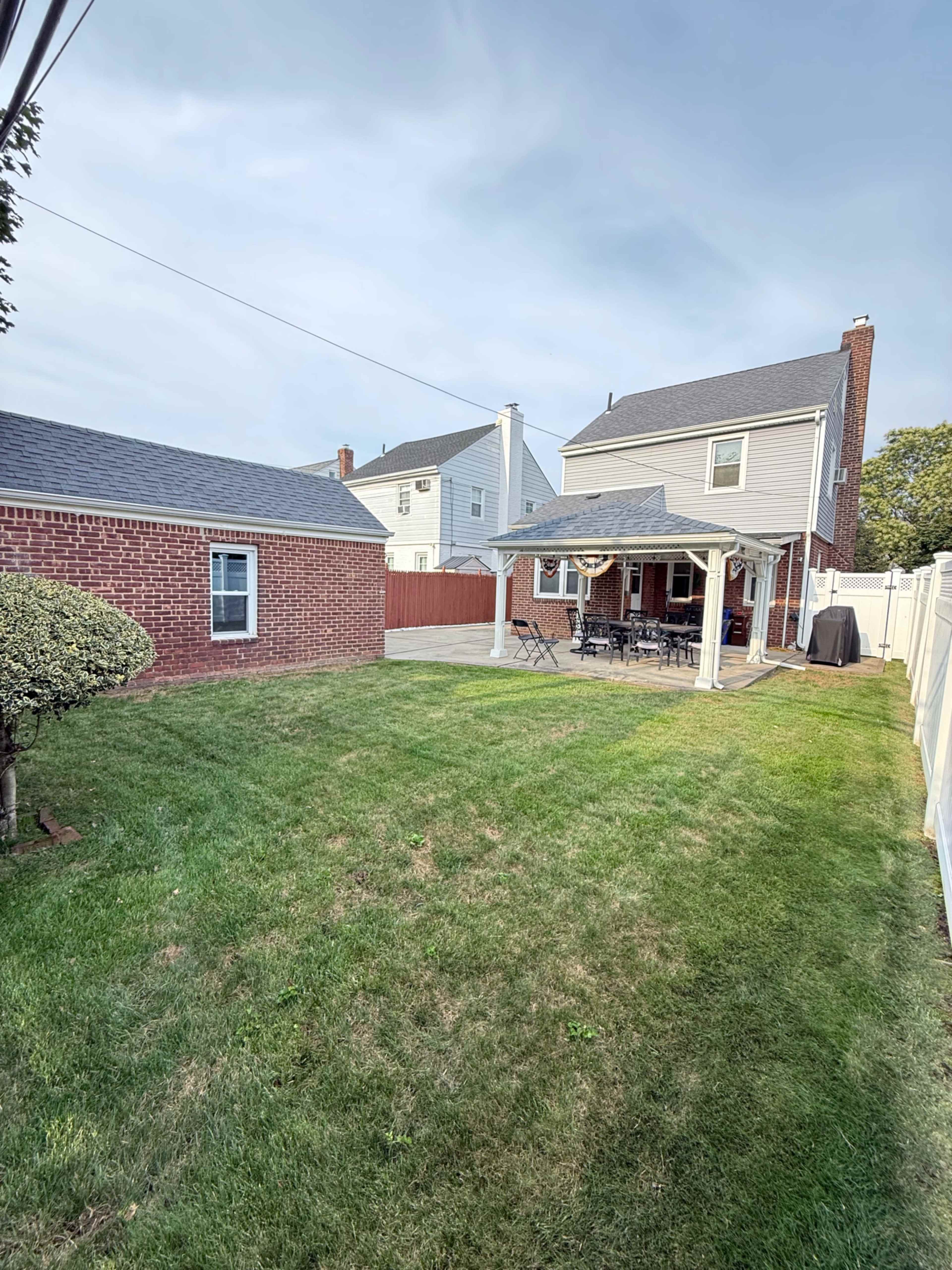 The image shows a grassy backyard with a patio area, a covered seating space, and two houses in the background.