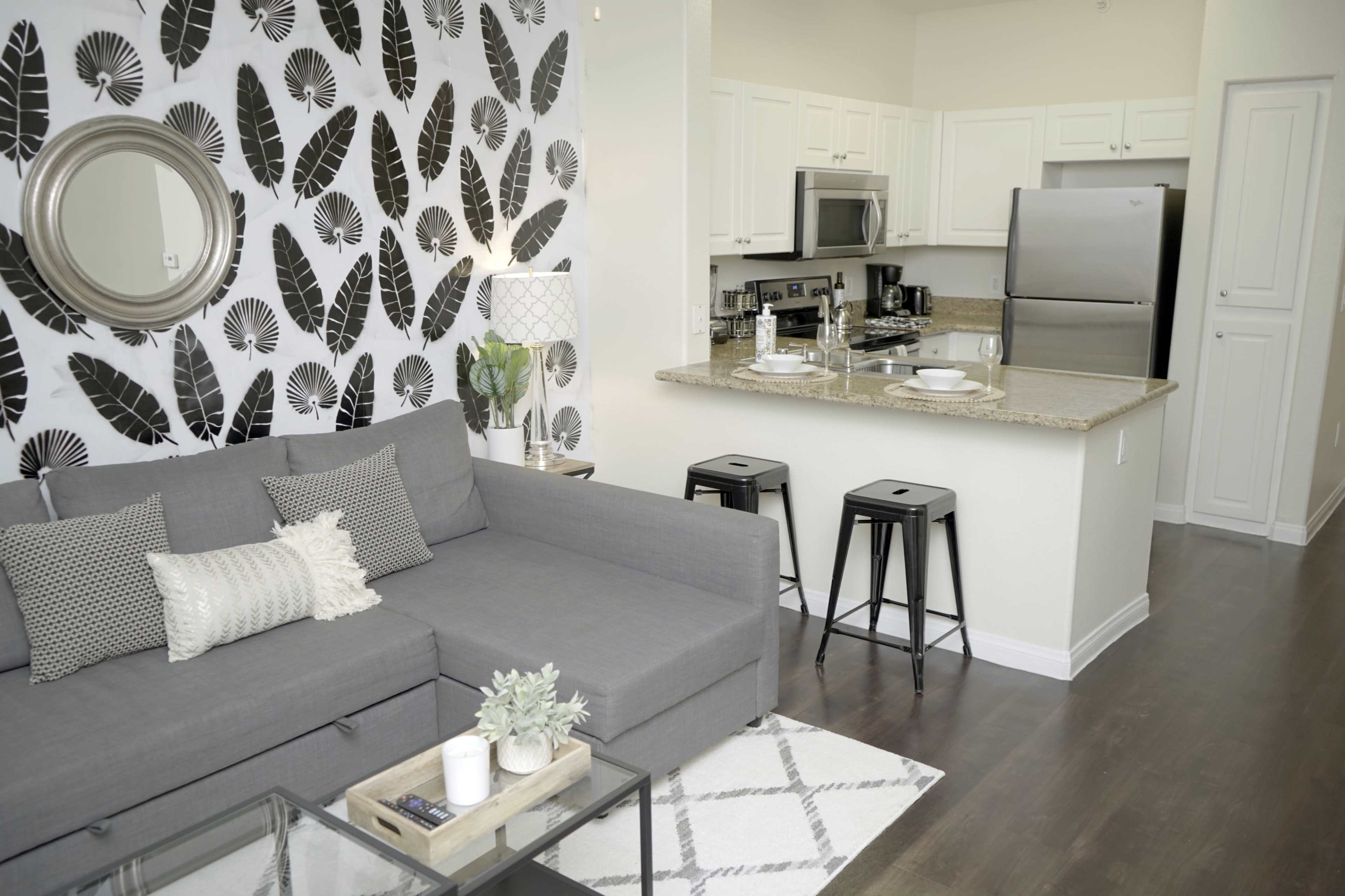 The image depicts a modern living space featuring a gray sectional sofa, a patterned accent wall, a kitchen with stainless steel appliances, and bar stools along the counter.
