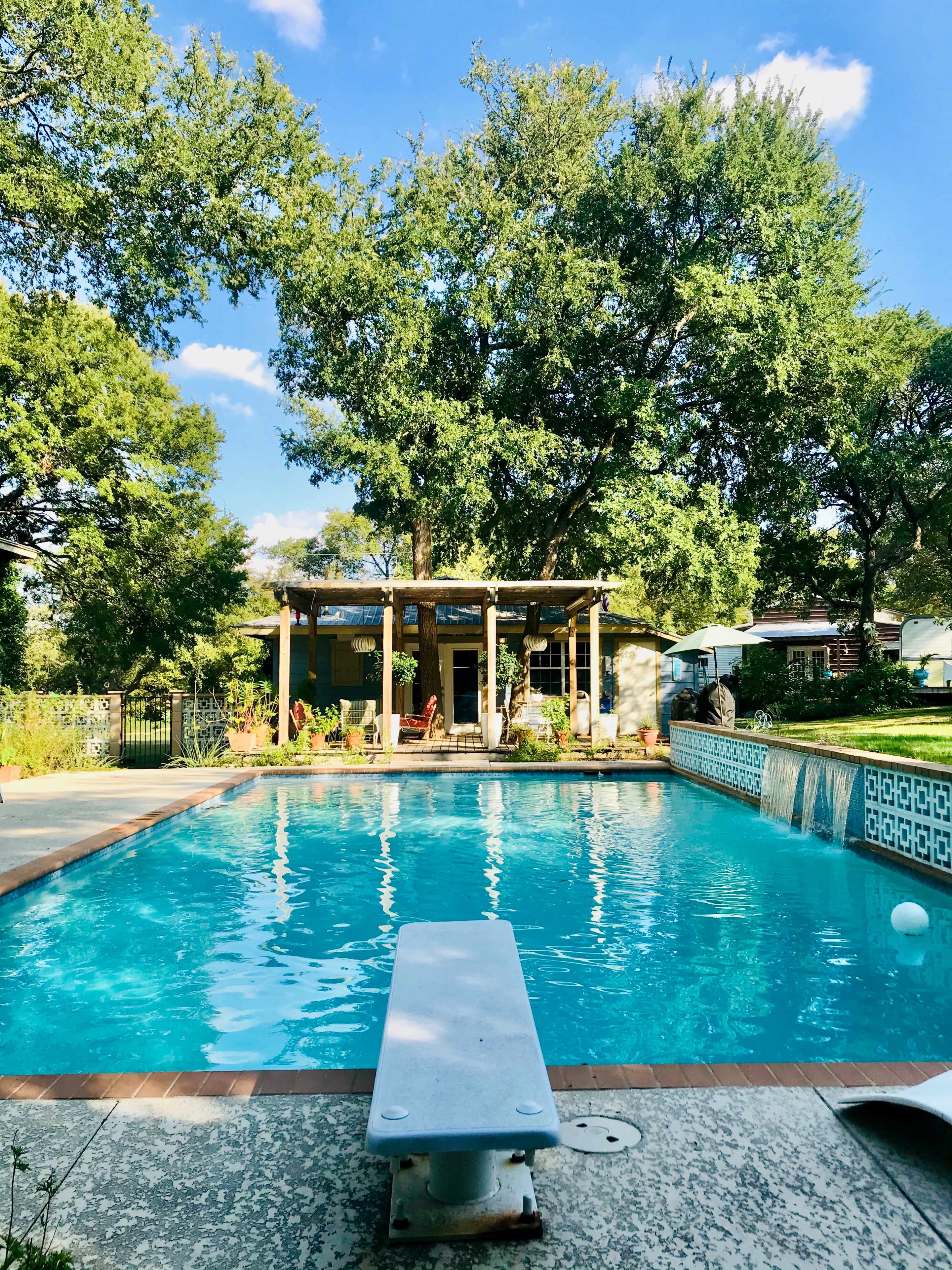 A clear swimming pool with a diving board is surrounded by lush greenery and a patio with a pergola in the background.