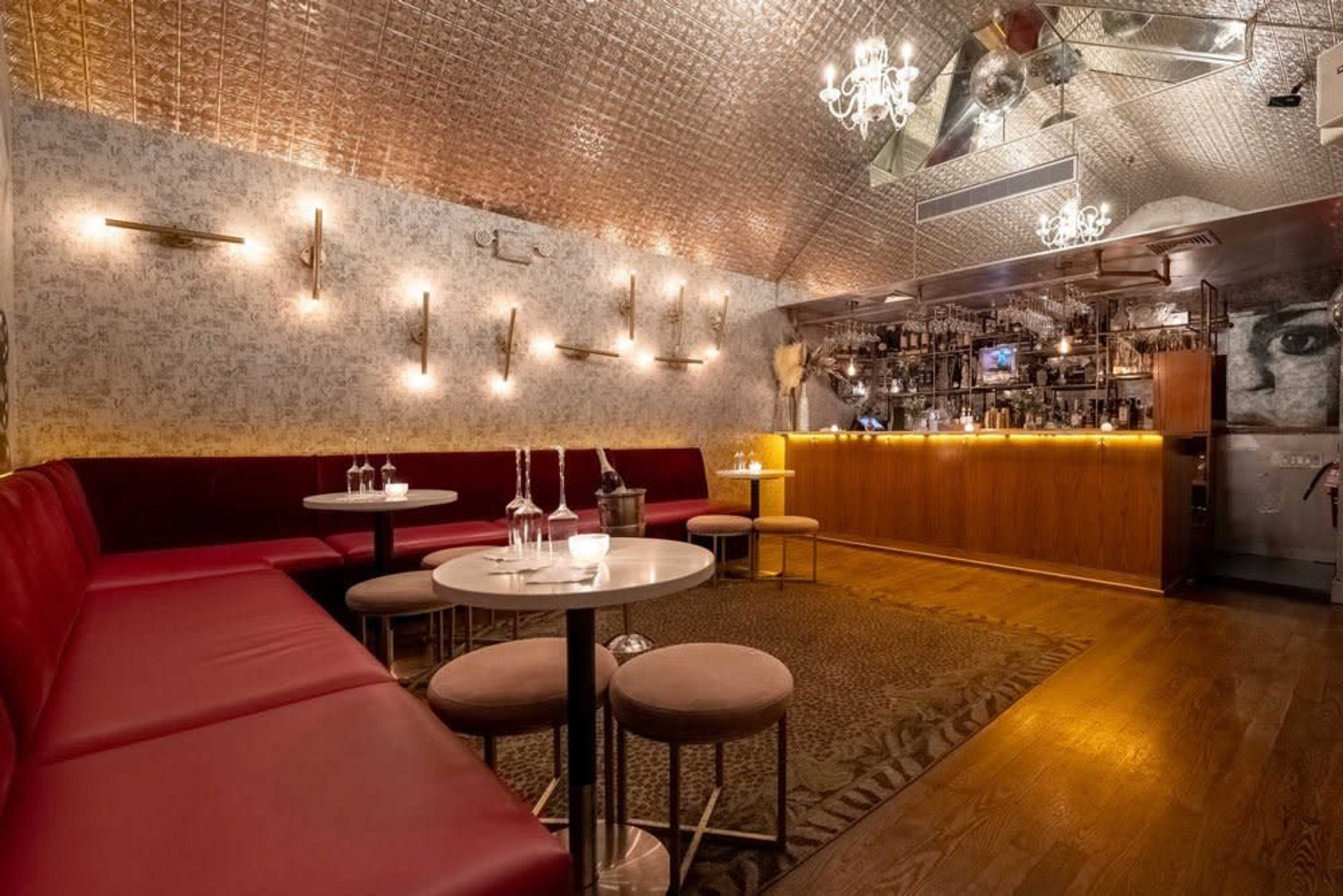The image depicts a modern, intimate bar setting with red upholstered seating, round tables, and an illuminated bar area in the background.