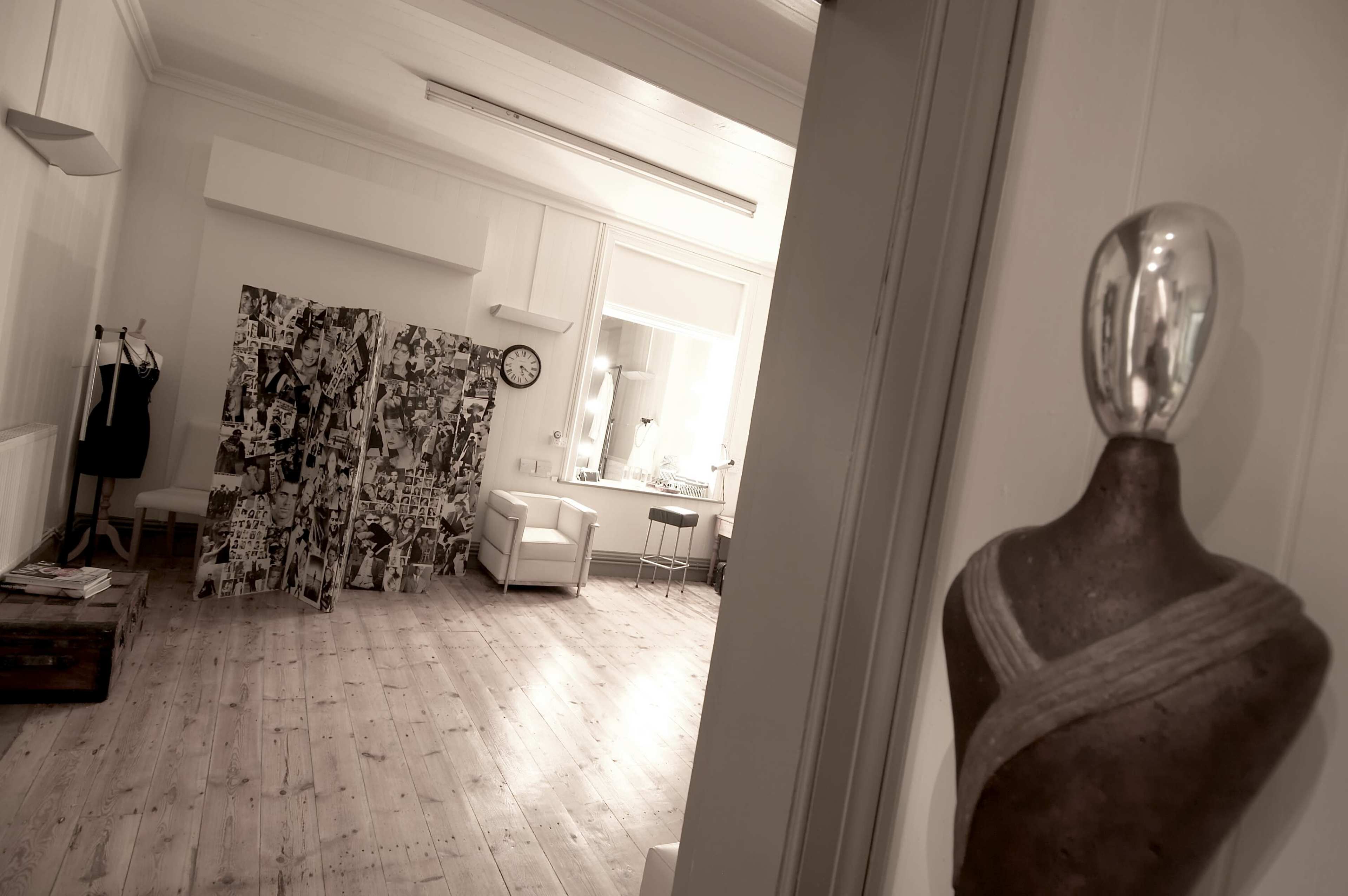 A spacious, minimalist room with a wooden floor, a large decorative screen made of collaged images, and modern furniture including a white chair and a wall clock.