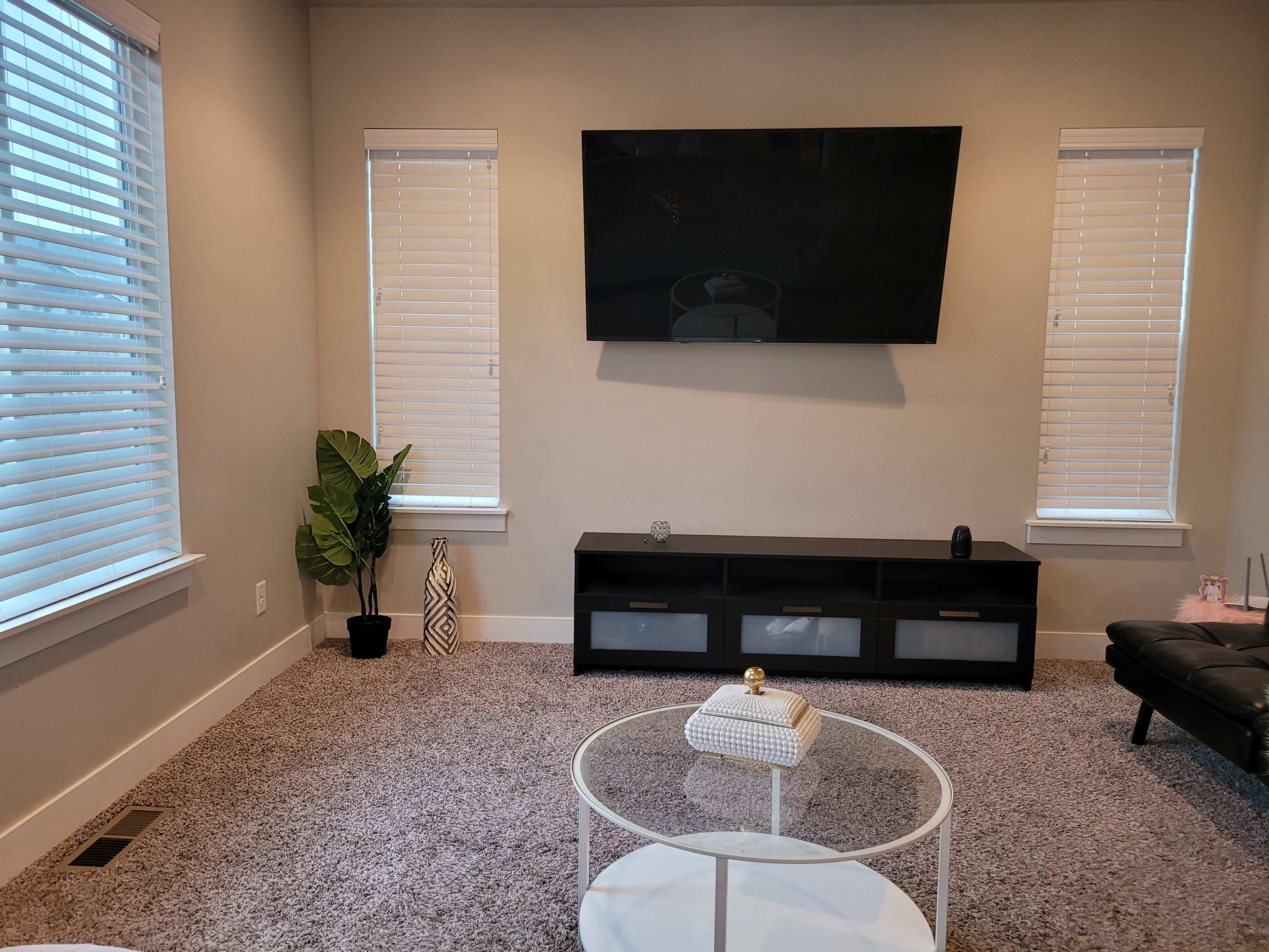 A living room featuring a television mounted on the wall, a black media console with drawers, a round coffee table, and two windows with blinds.