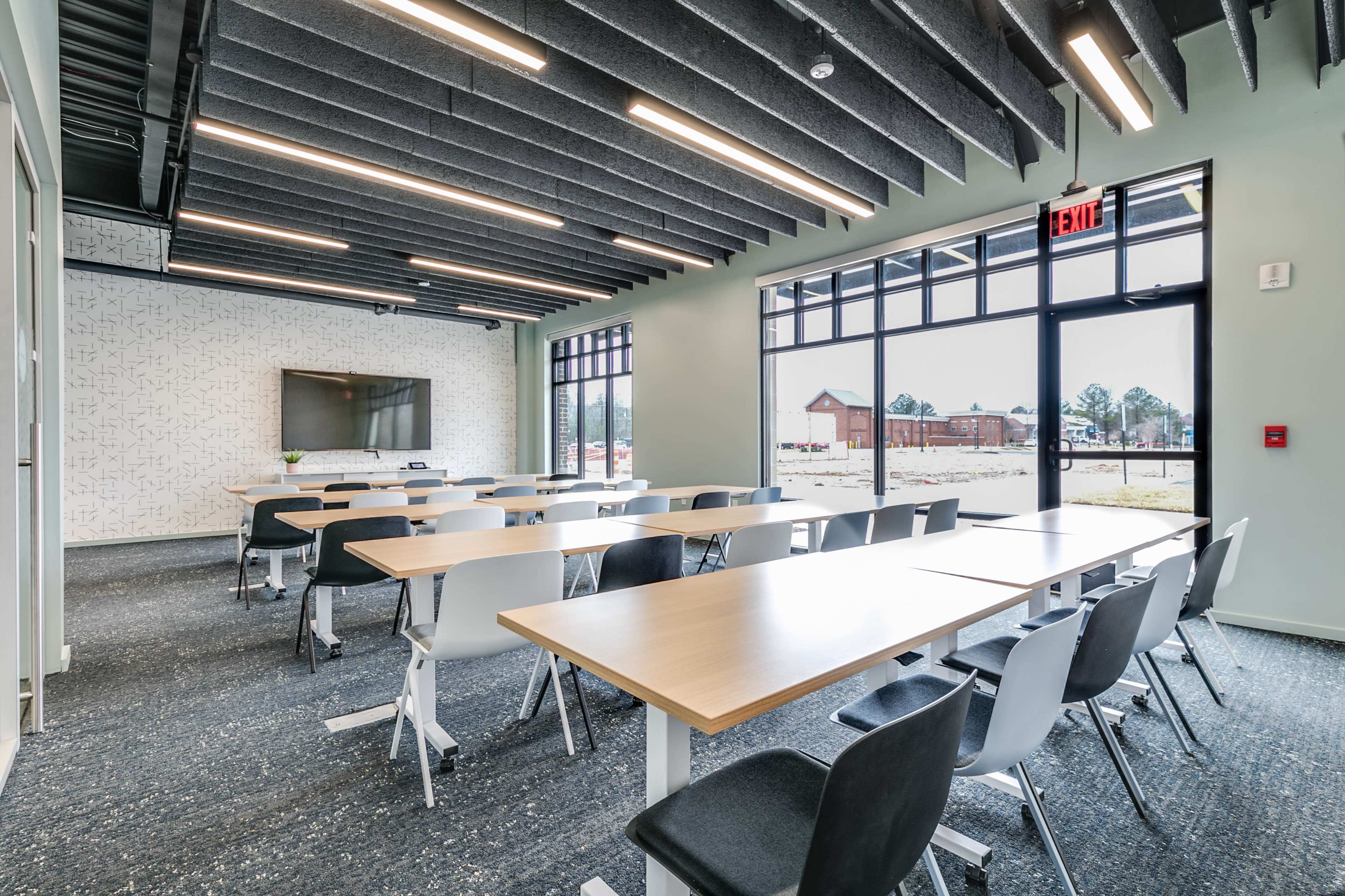 A modern conference room with rows of tables and chairs, large windows providing natural light, and a wall-mounted screen for presentations.