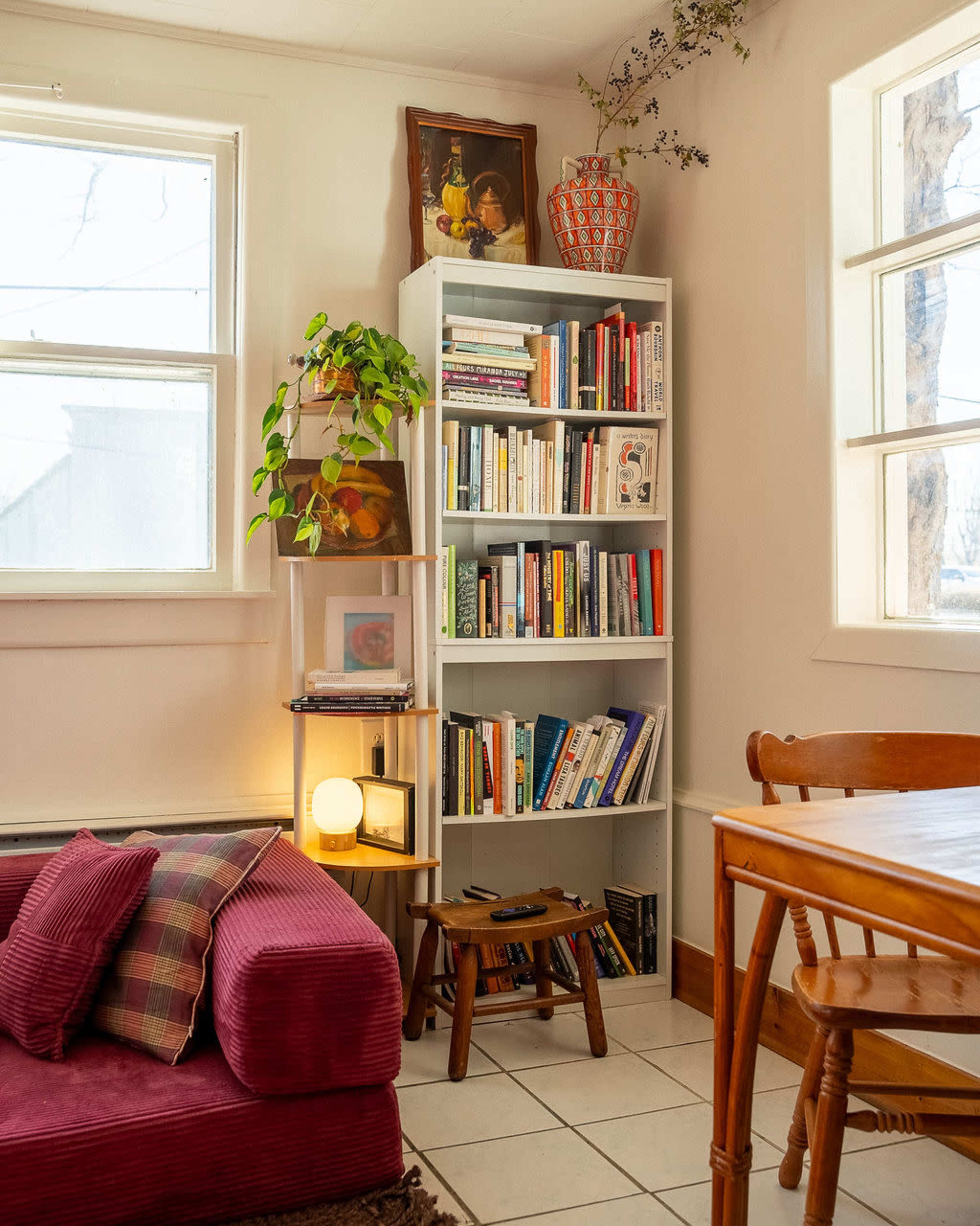 The scene shows a cozy living space with a bookshelf filled with books, a small wooden table and chairs, and a sofa adorned with cushions.