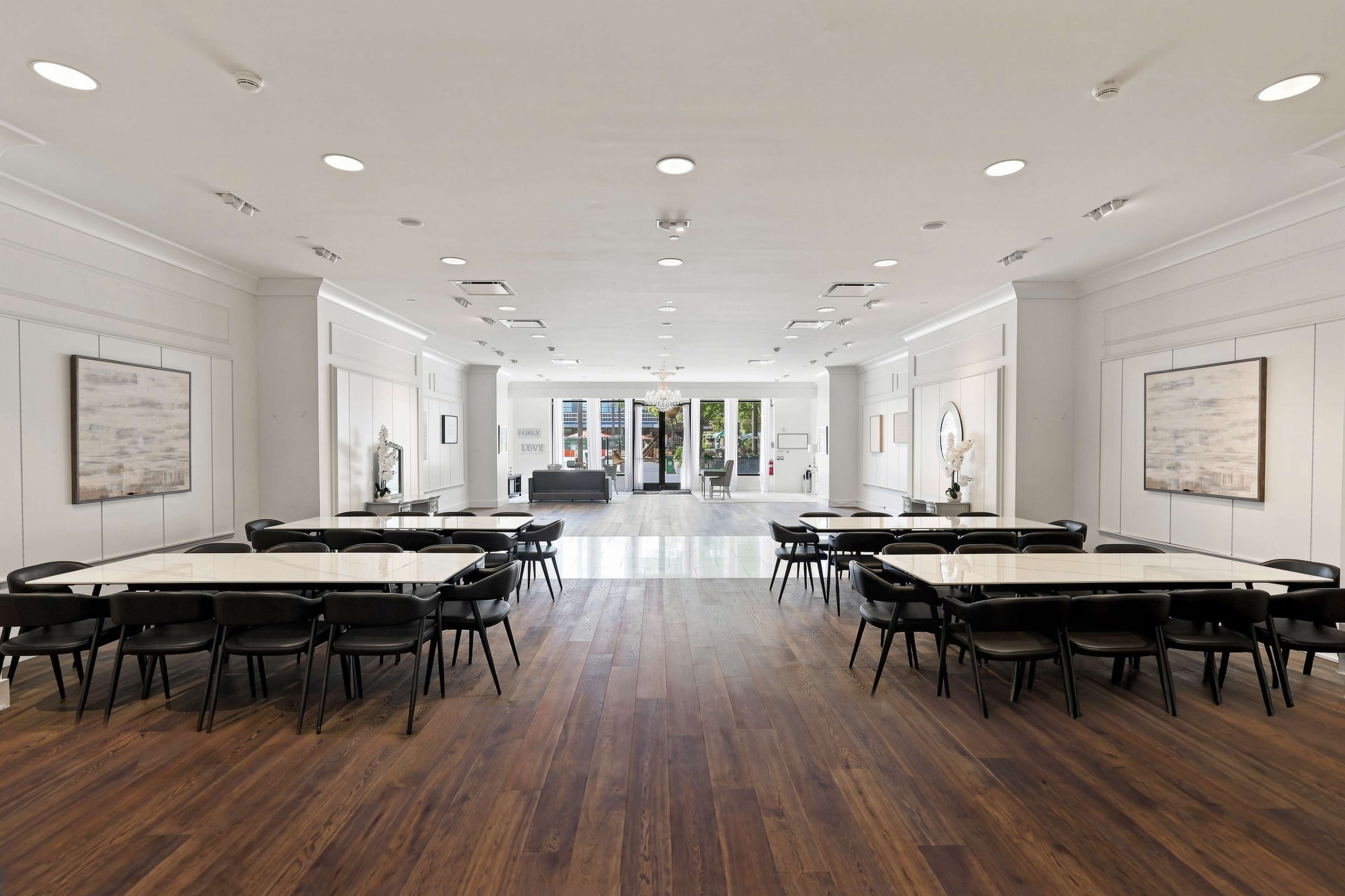 The image shows a spacious event room with several long tables arranged in two rows, featuring hardwood flooring and large windows in the background.
