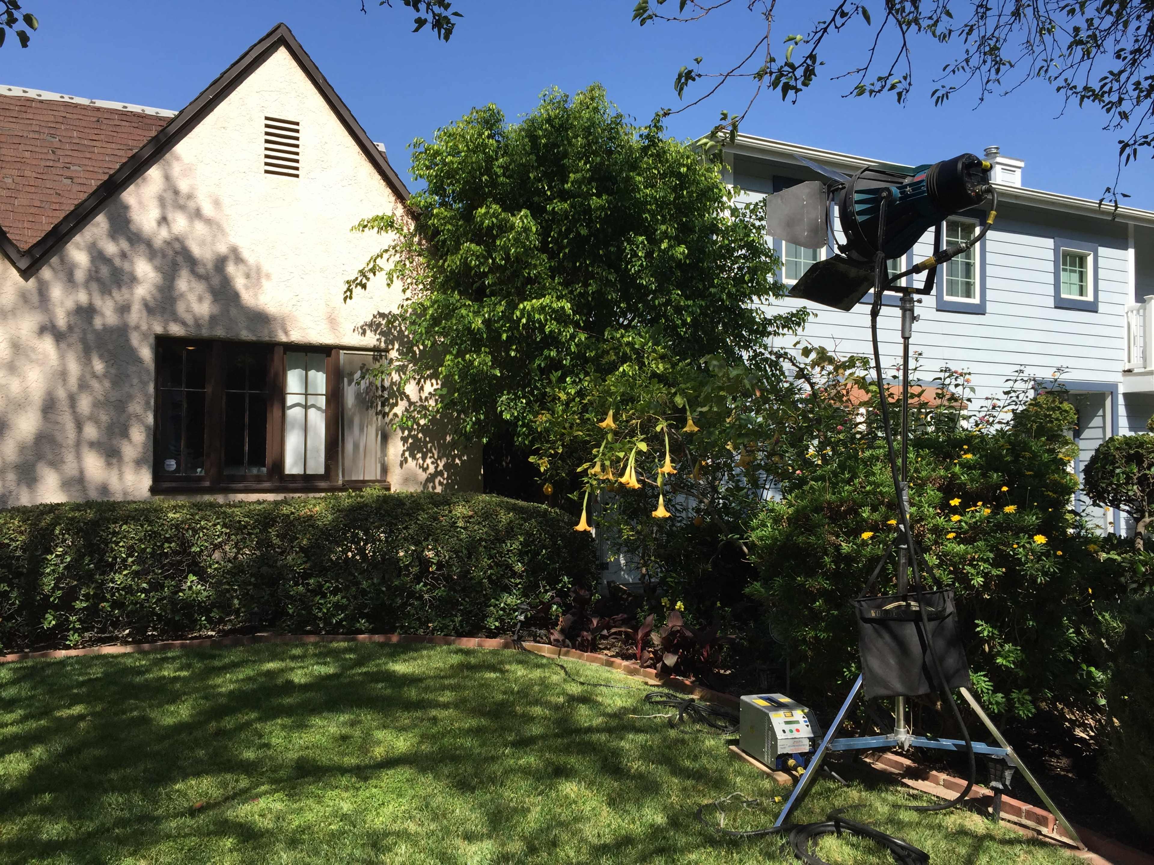 A bright backyard scene features a house with a cream-colored exterior and a green lawn, alongside a film light mounted on a stand near a tree.