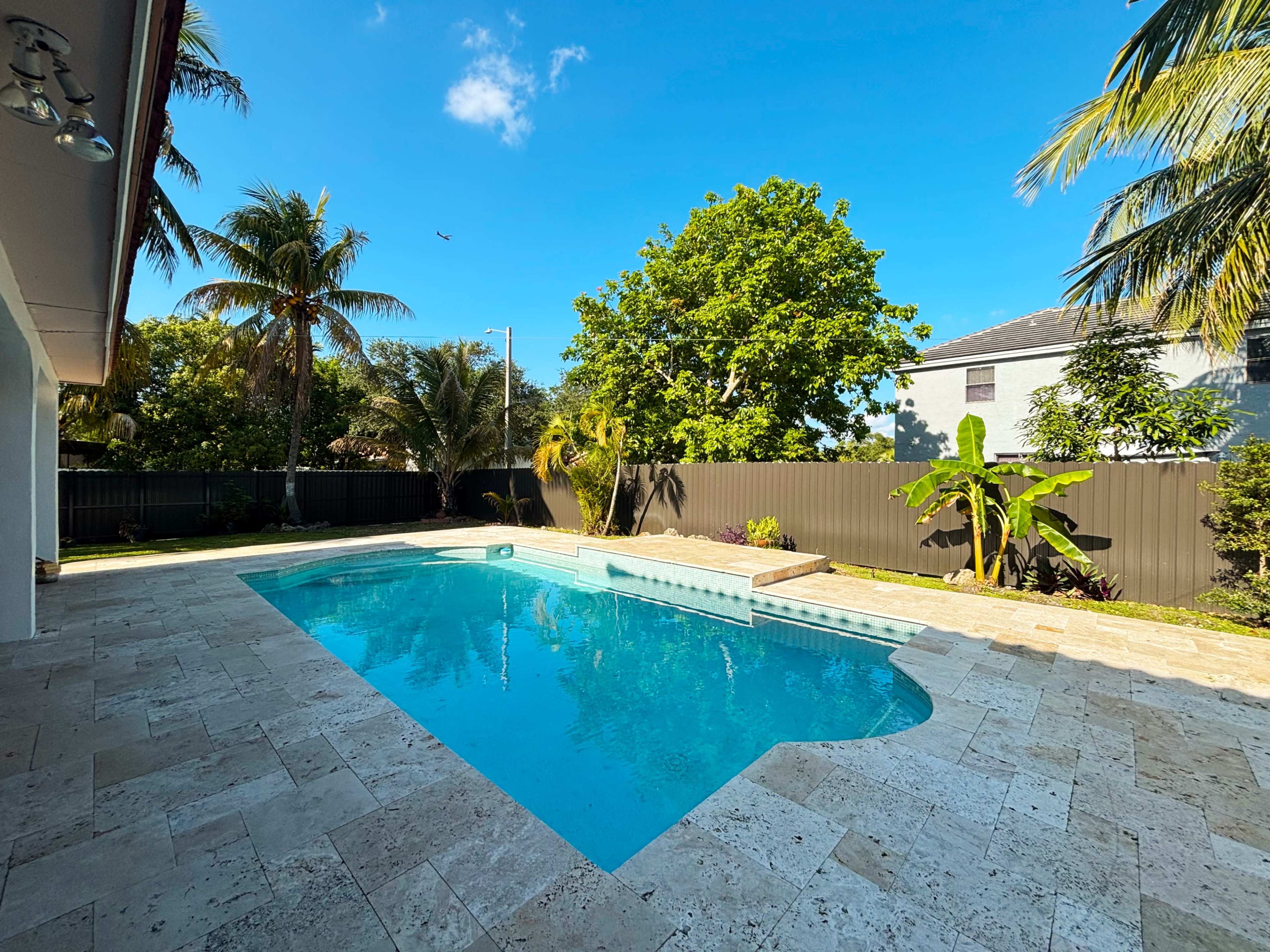 The image shows a landscaped backyard featuring a swimming pool surrounded by tropical plants and palm trees.