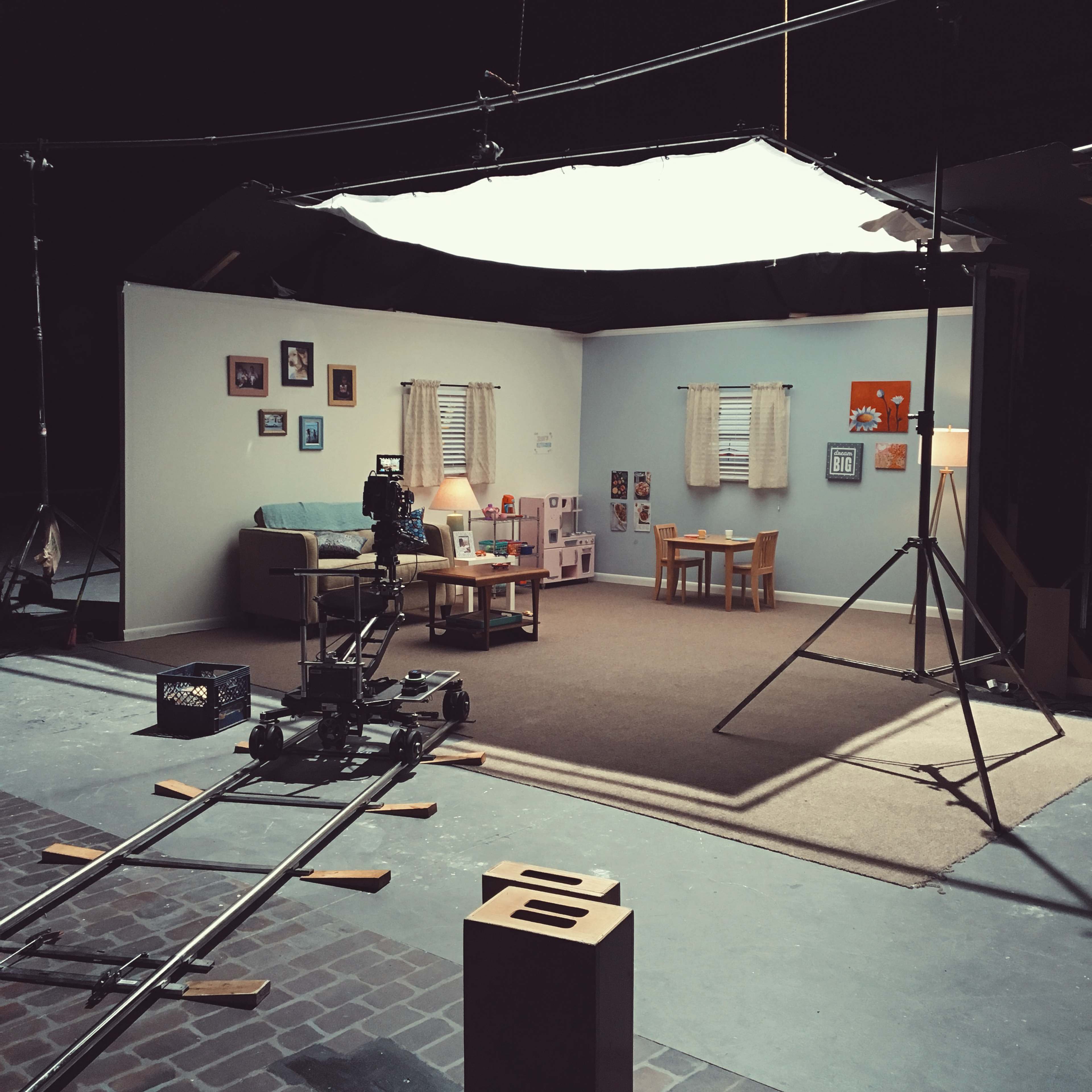 A film set features a cozy living room scene with a sofa, two chairs, children's toys, and a table arranged on a carpeted floor.