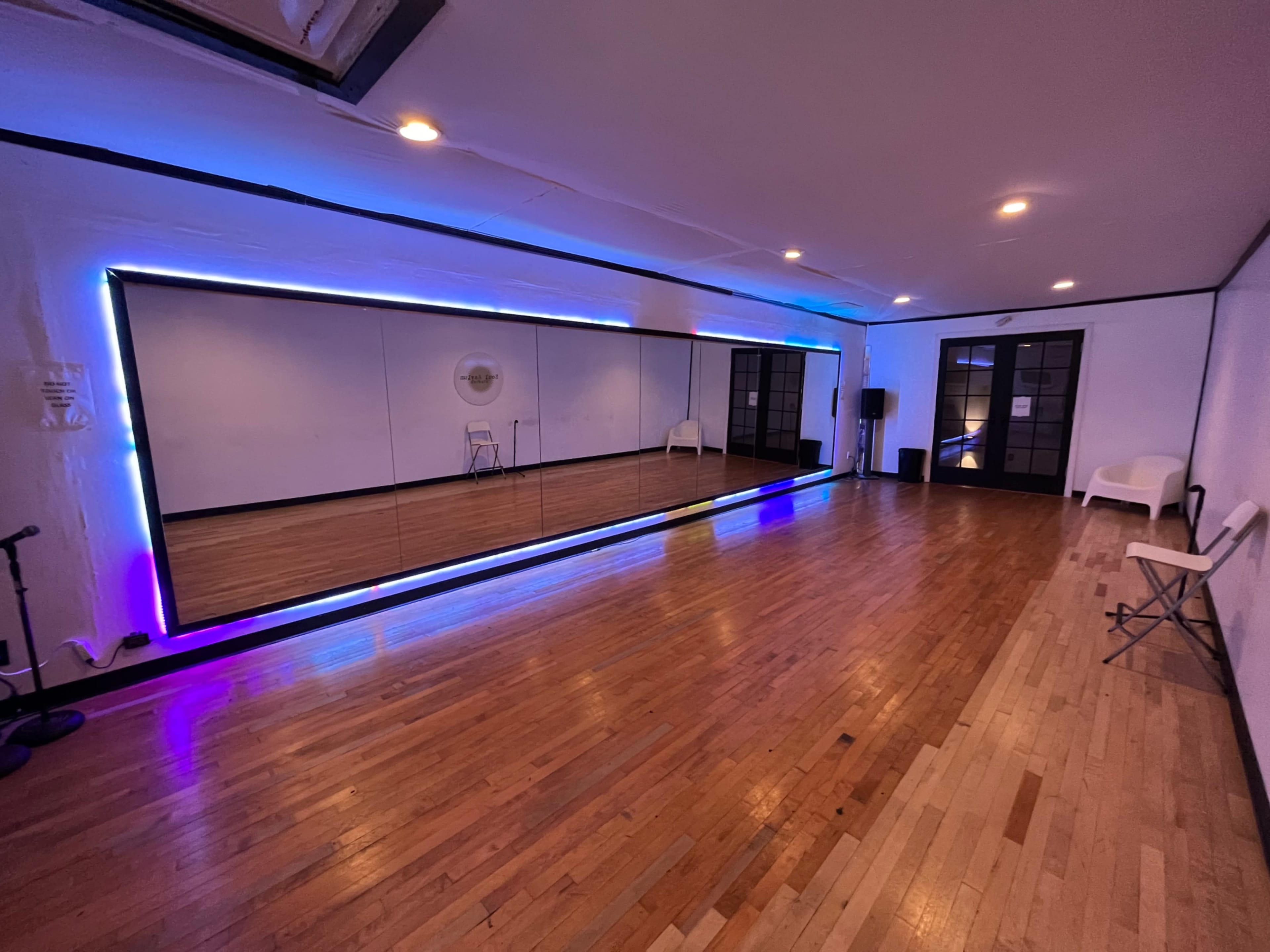 A spacious room with wooden floors, a large mirror framed by neon lights, and minimal seating along the walls.