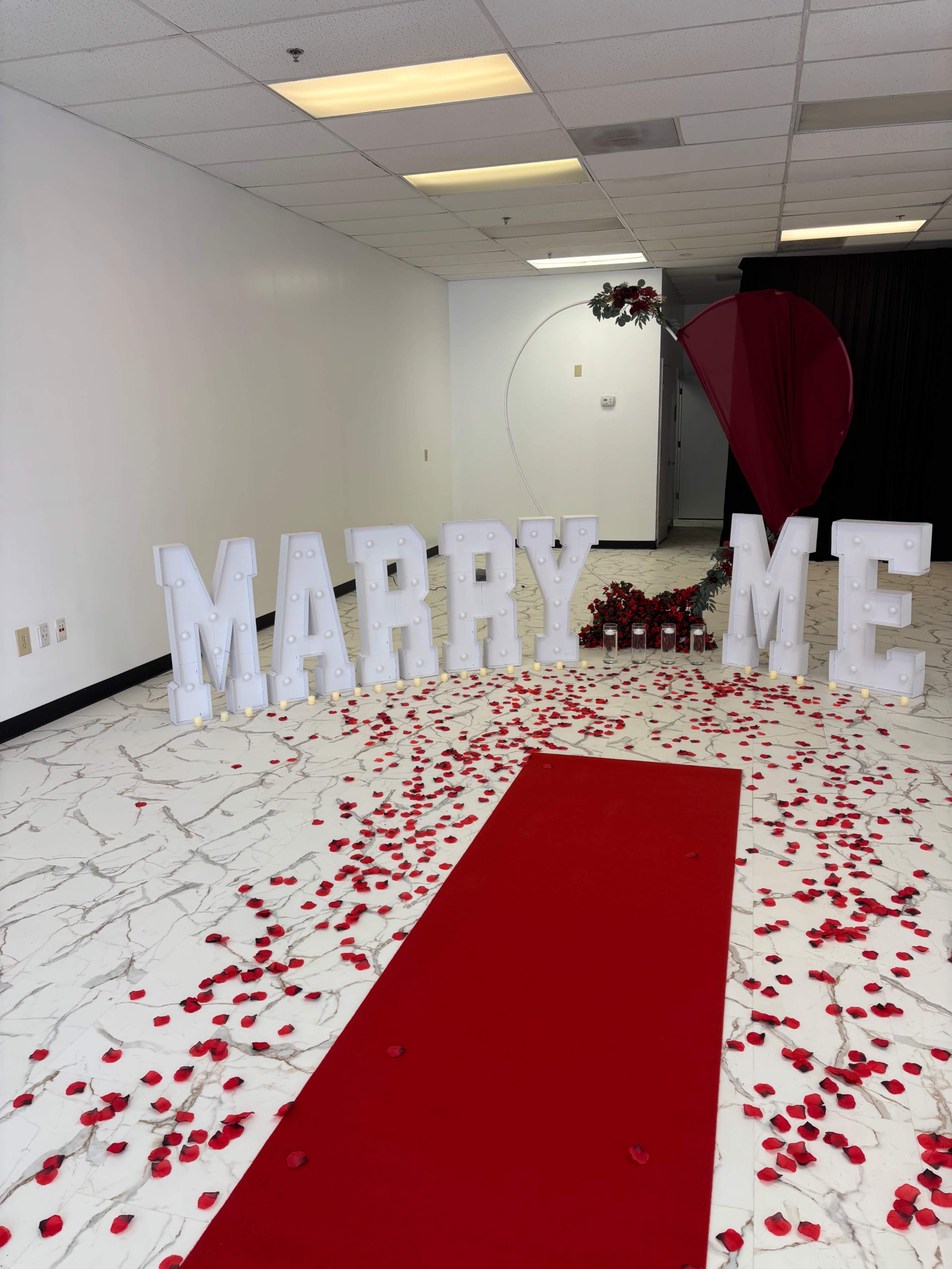The image shows a decorated space with large illuminated letters spelling "MARRY ME," surrounded by a red carpet and scattered rose petals.