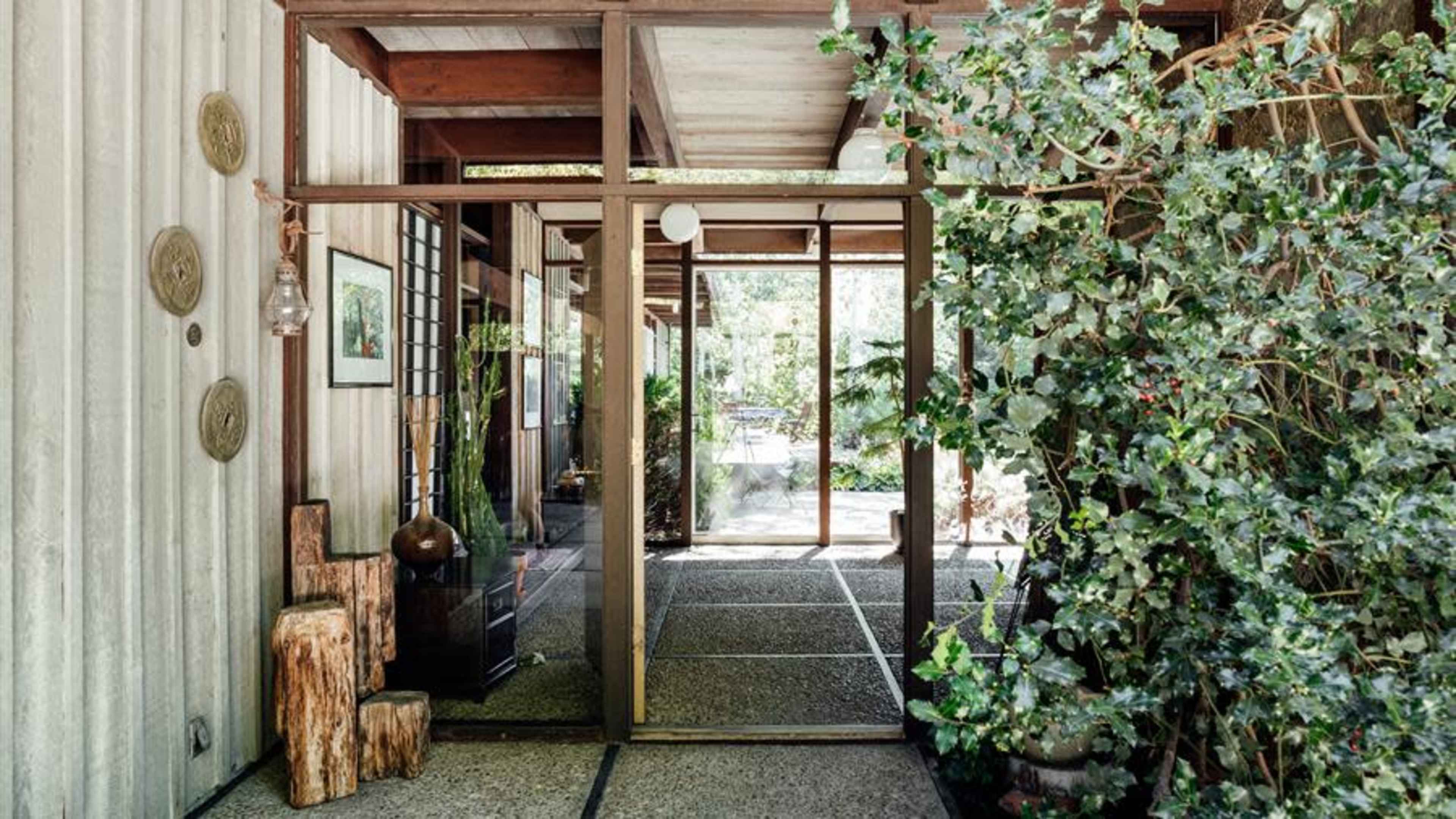 A modern entrance lined with concrete flooring, wooden beams, and greenery surrounding glass doors leading into a landscaped outdoor area.