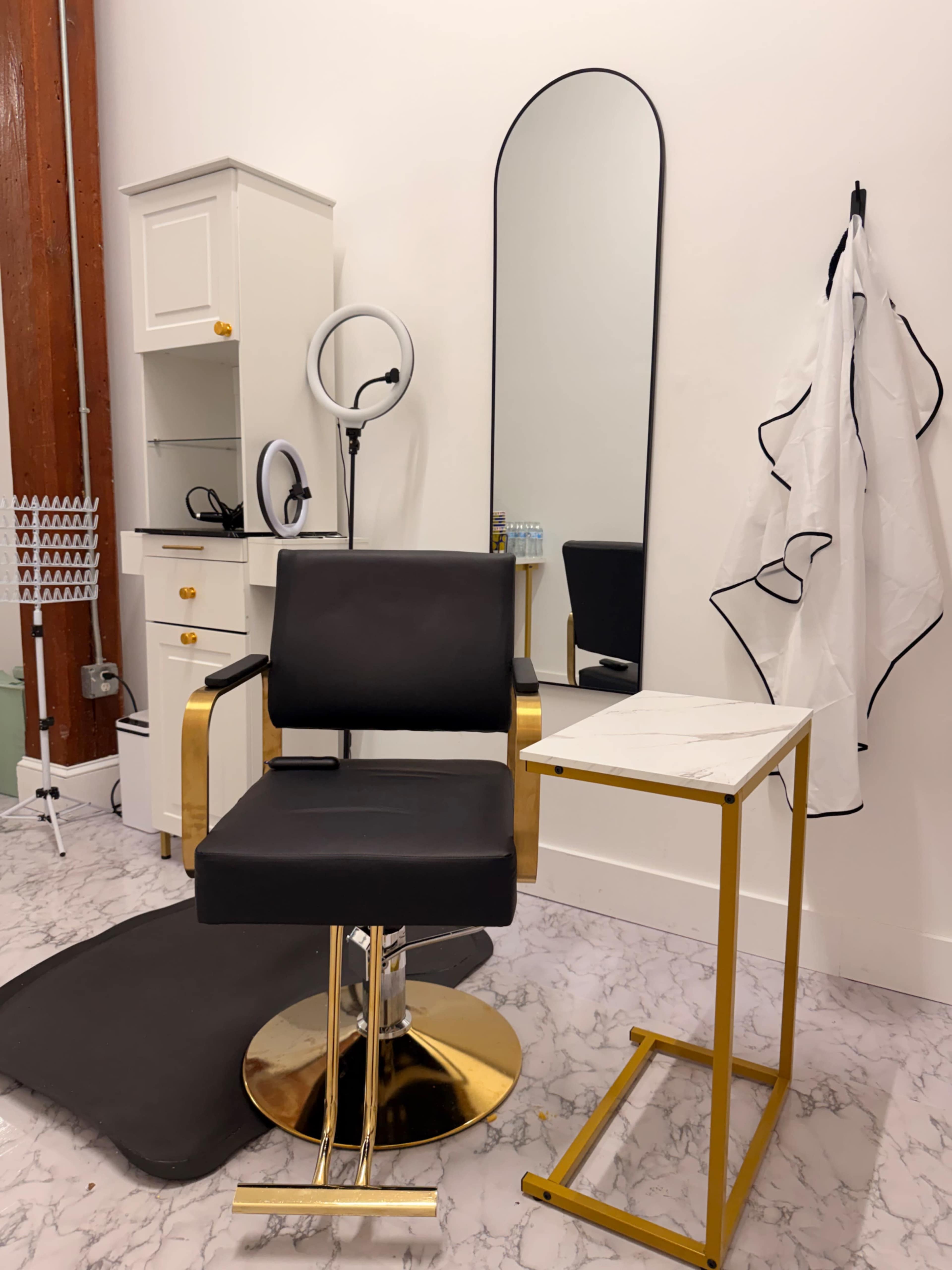 The image shows a stylish hair salon setup featuring a black salon chair, a small marble-topped table, a large mirror, and a ring light.