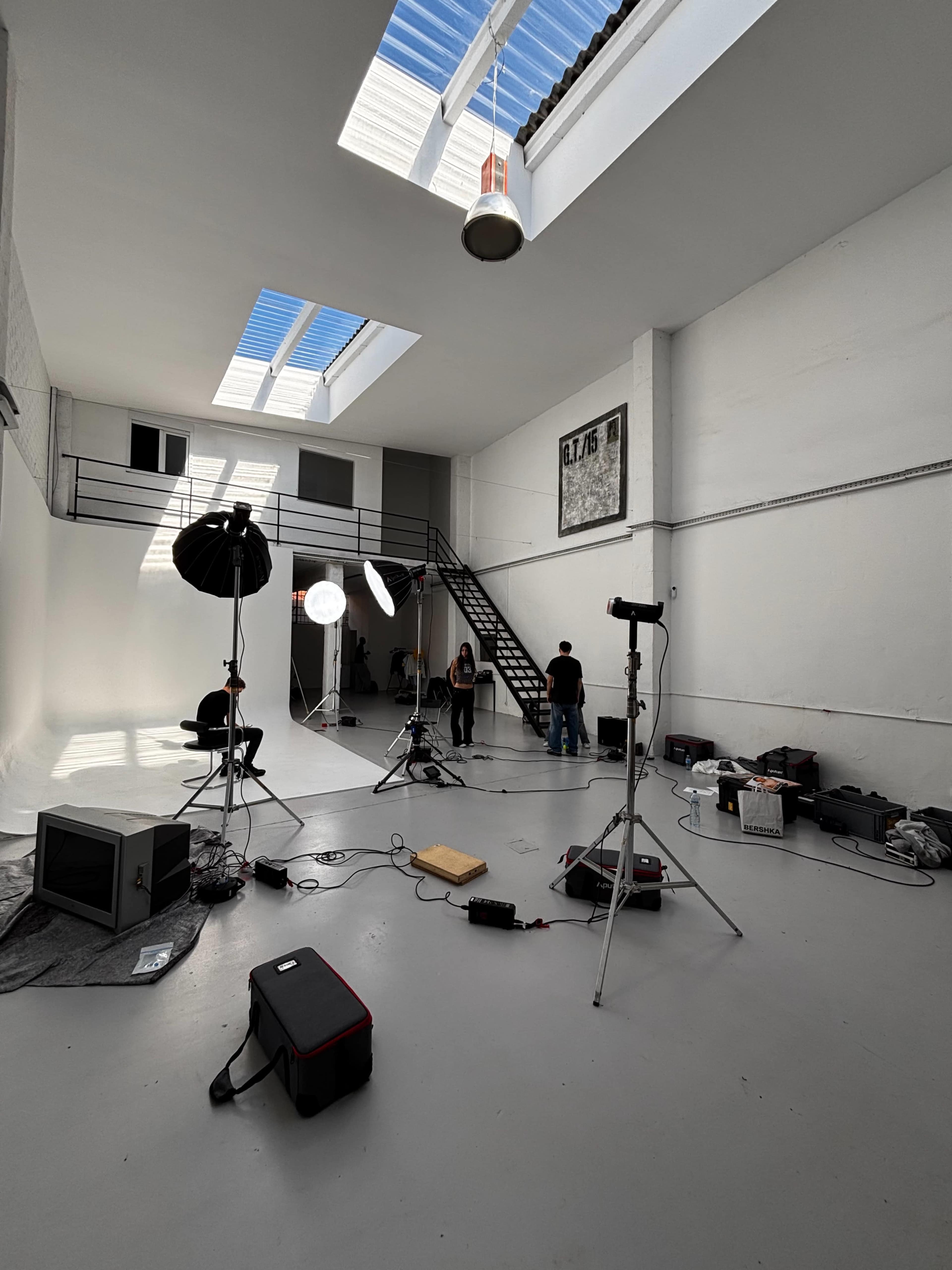 The image shows a spacious photography studio with multiple lighting setups, equipment scattered across the floor, and a staircase visible on the far side.