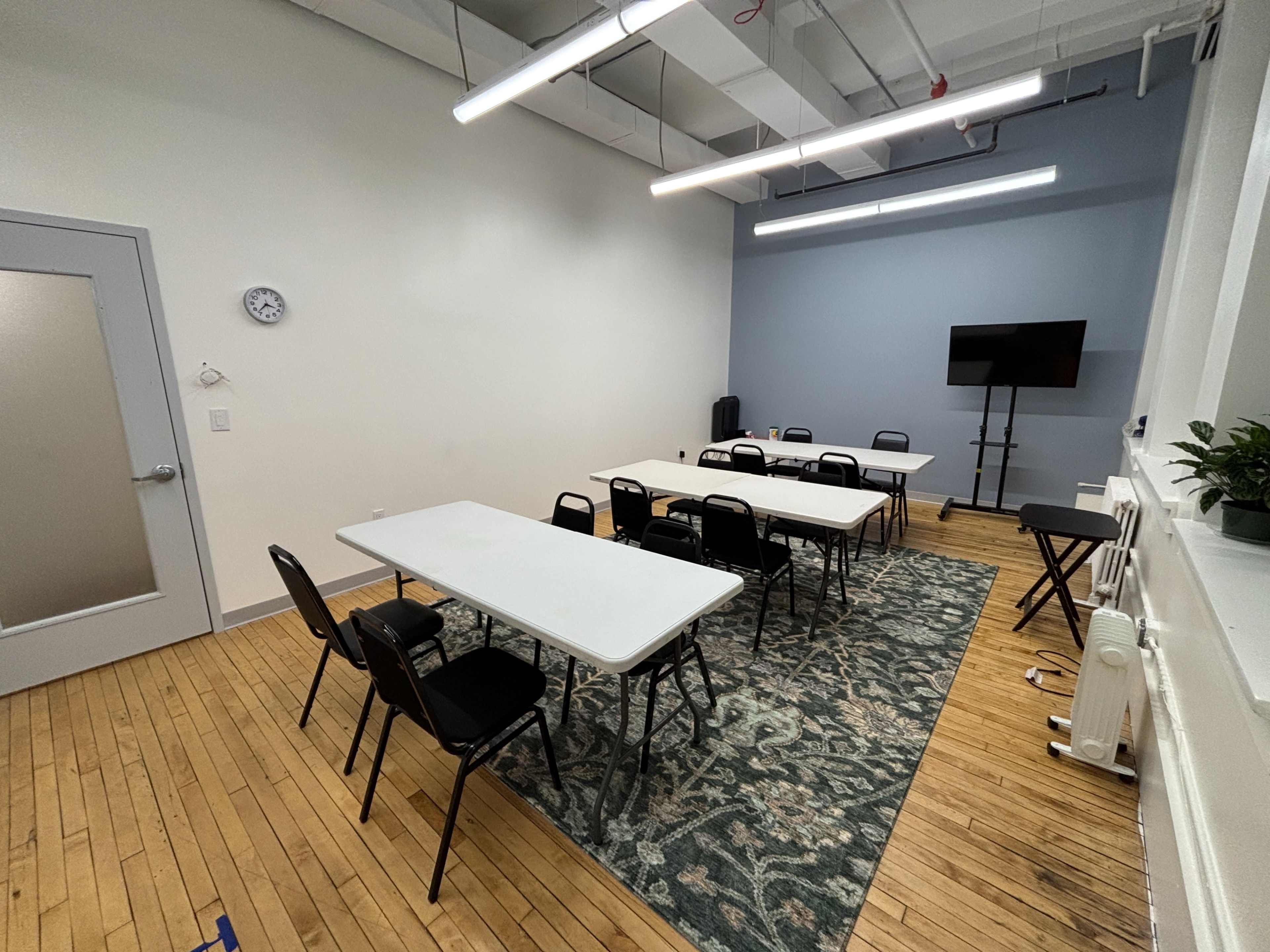 The image shows a spacious meeting room with multiple tables arranged in a U-shape, surrounded by black chairs, and featuring a television on a stand and a potted plant in the corner.