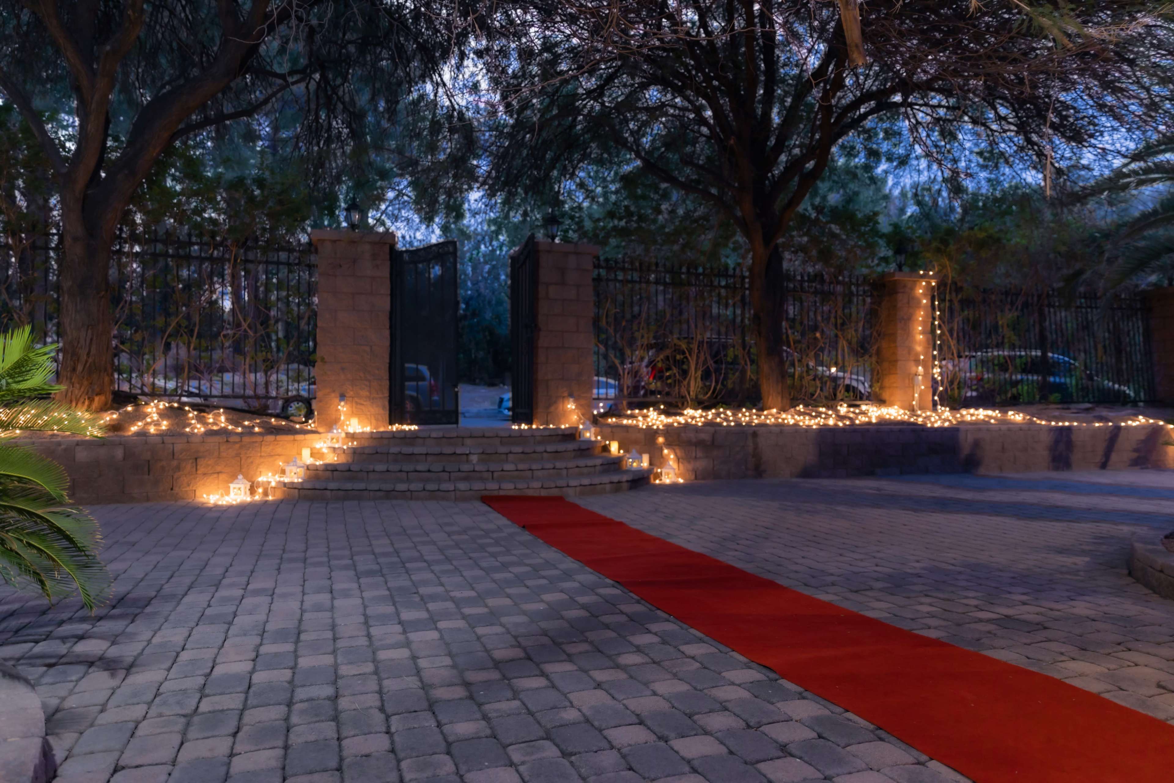 The scene features a pathway lined with glowing candles leading up to an ornate gate, surrounded by trees and illuminated by a soft evening light.