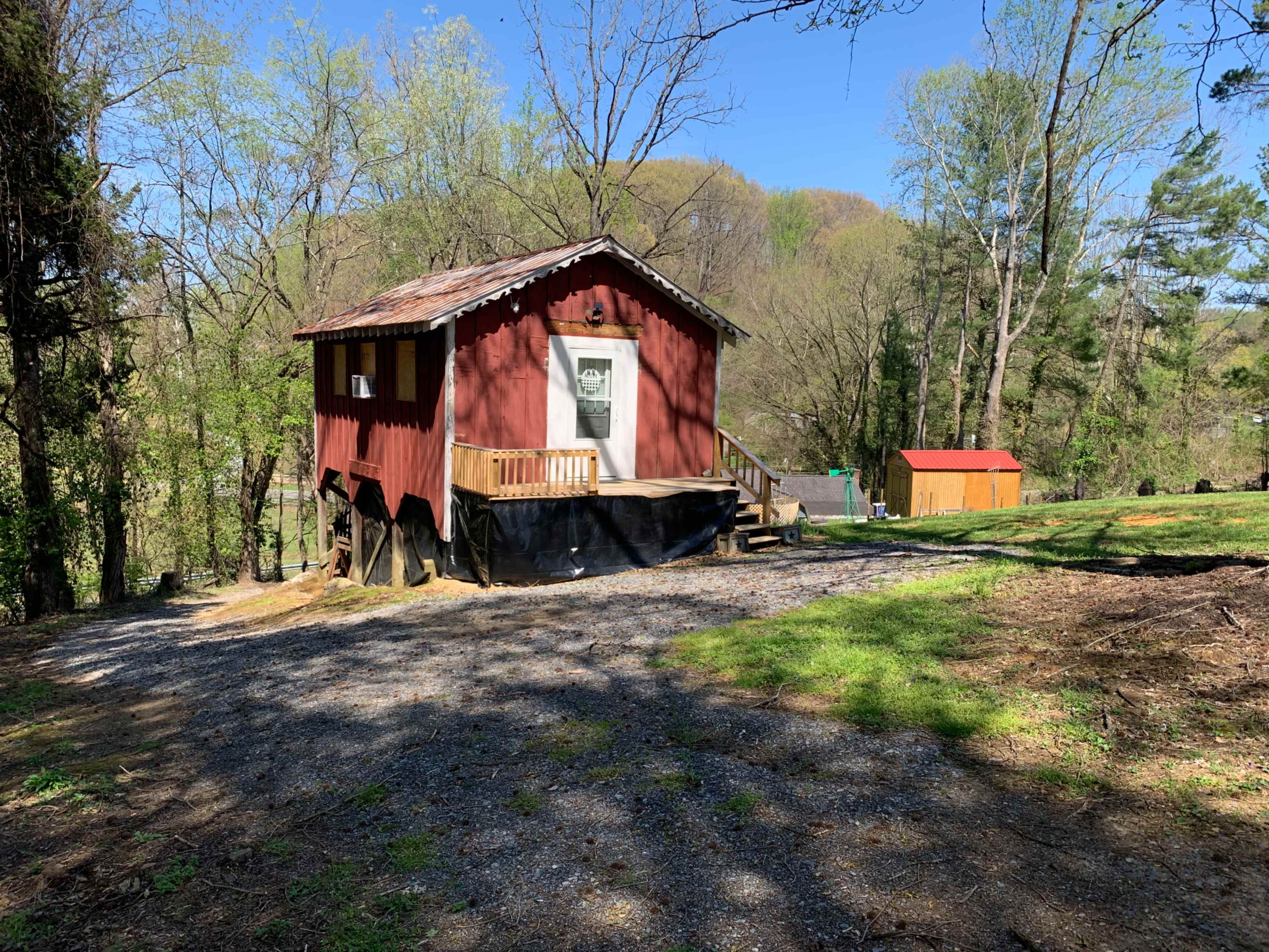 A small red house is situated on a gravel path surrounded by trees, with a covered porch and a storage shed in the background.