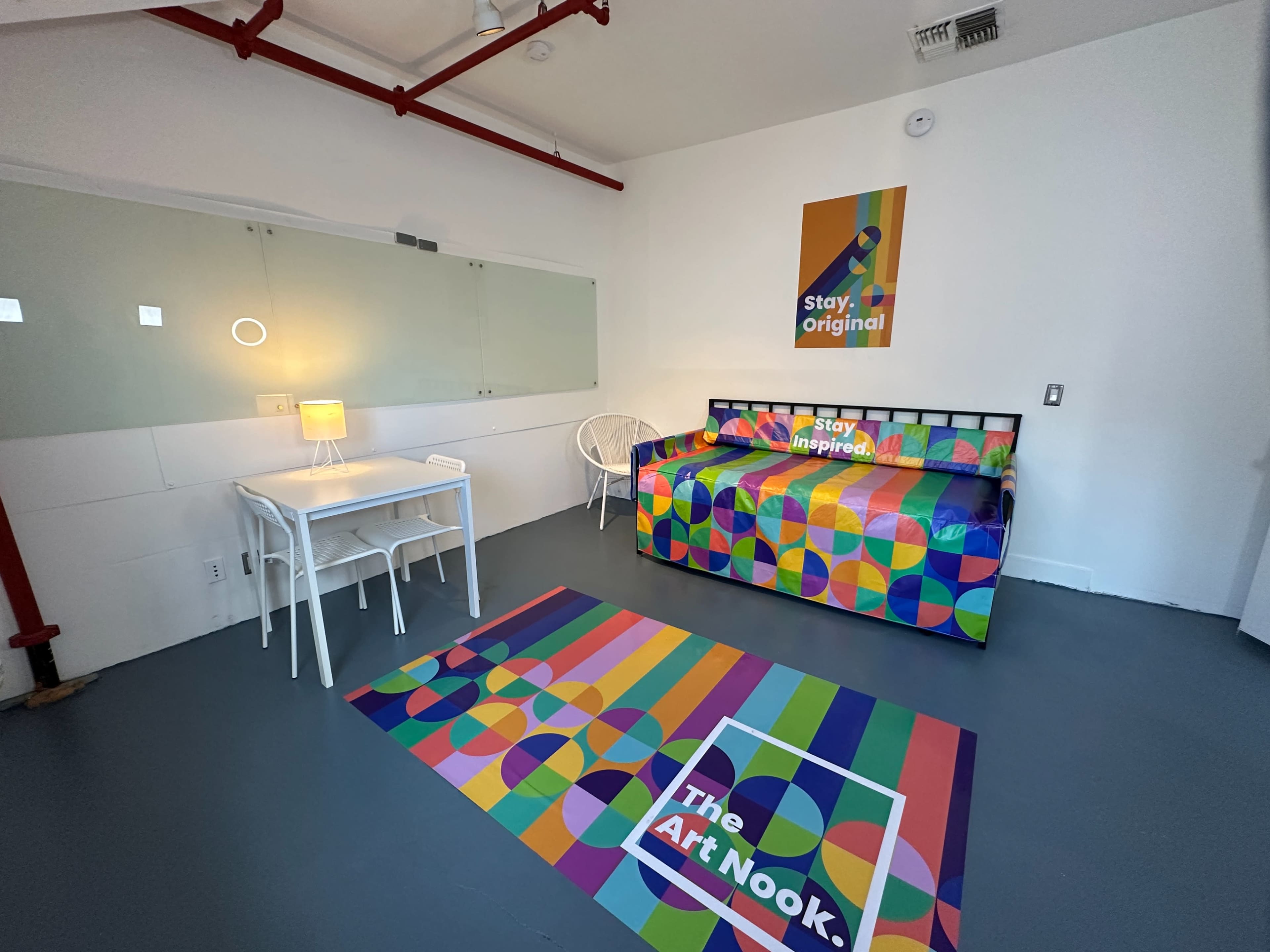 The image shows a brightly colored, modern room with a patterned daybed, a small white table with a lamp, and a large rug, all set against a minimalist white wall.