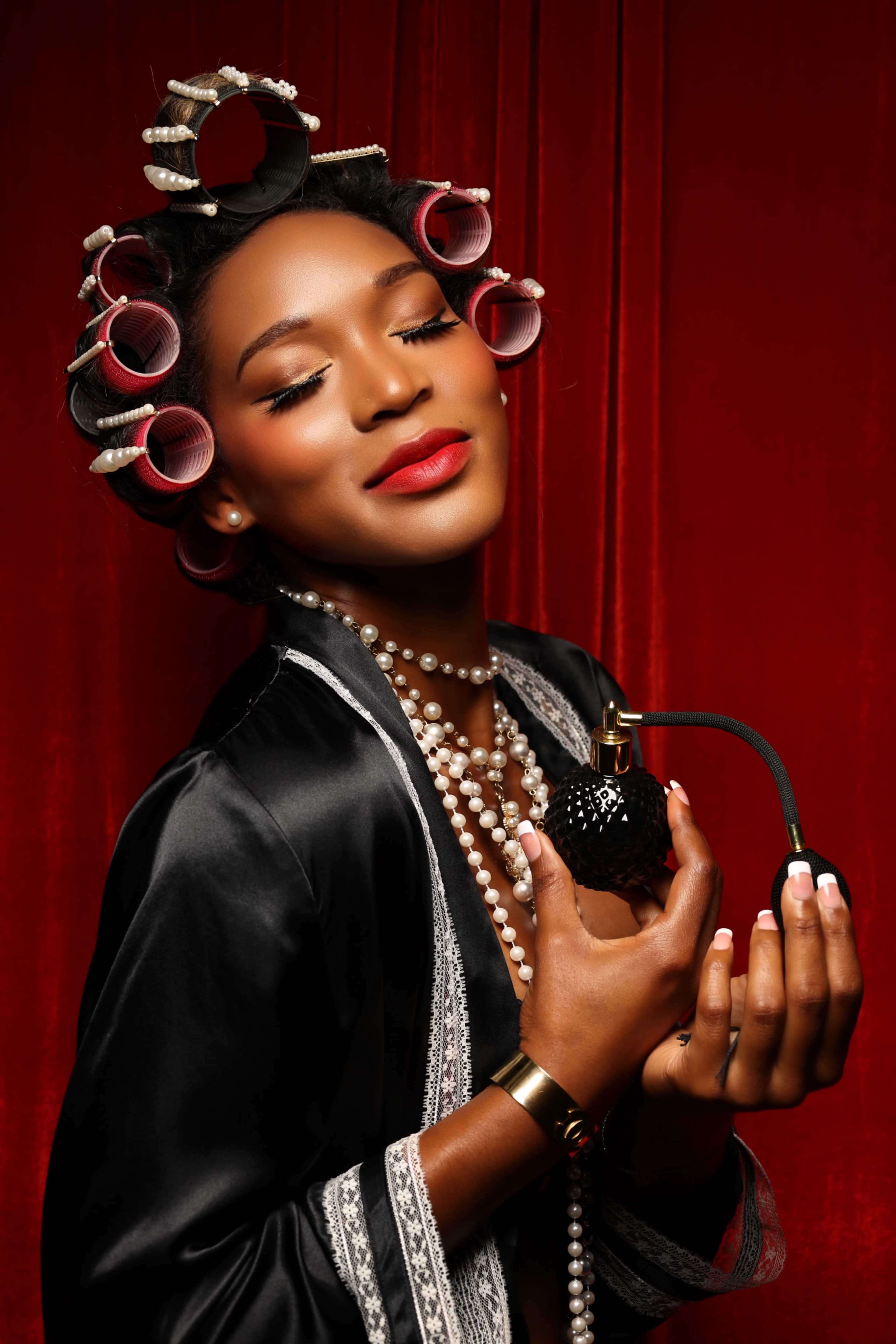 A woman in hair rollers holds a vintage perfume bottle, wearing a black robe and layered pearl necklaces against a red backdrop.