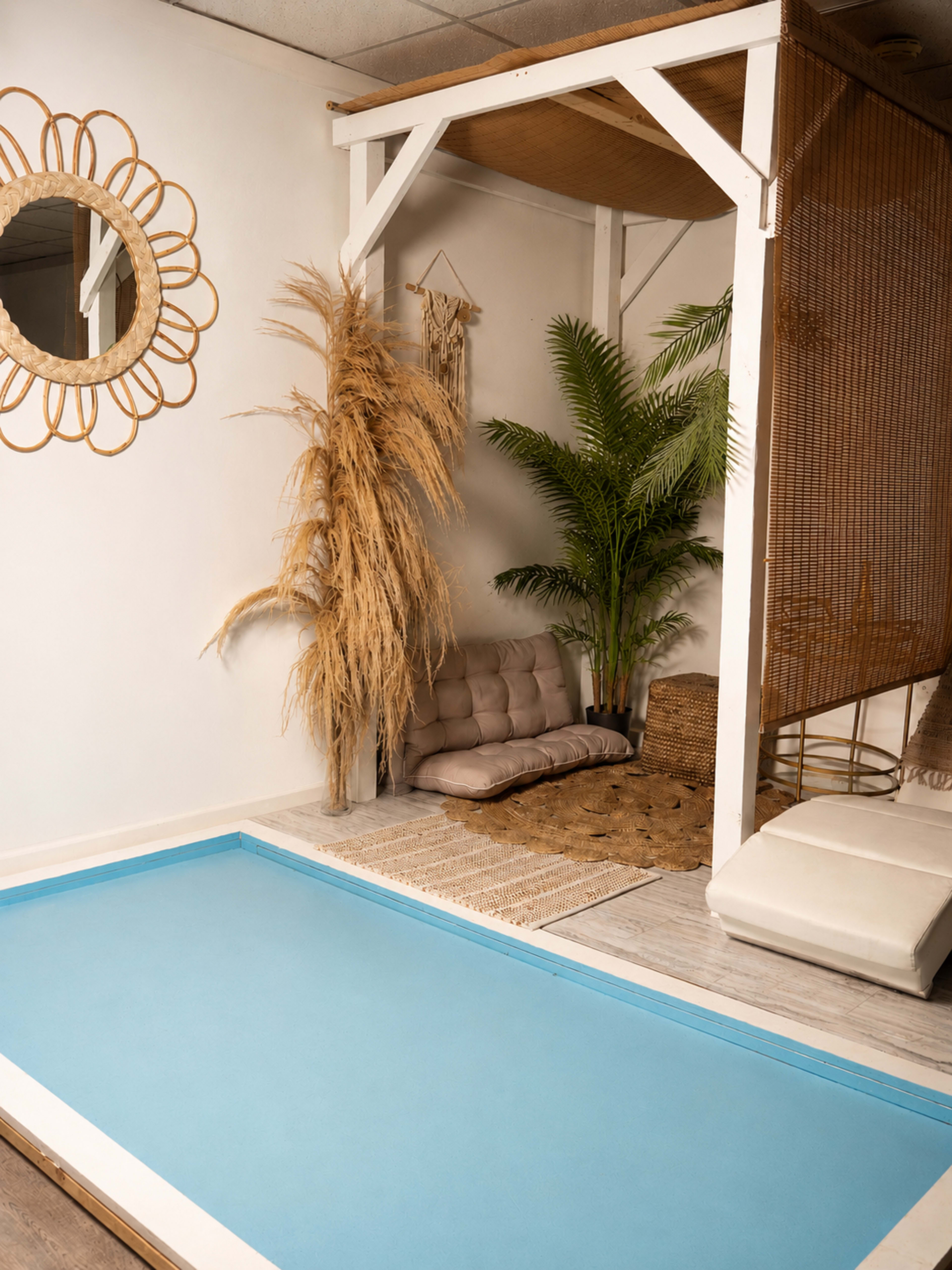 The image shows a modern indoor space featuring a blue mat, a rattan mirror, a potted plant, a couch, and decorative elements like woven baskets and dried foliage.