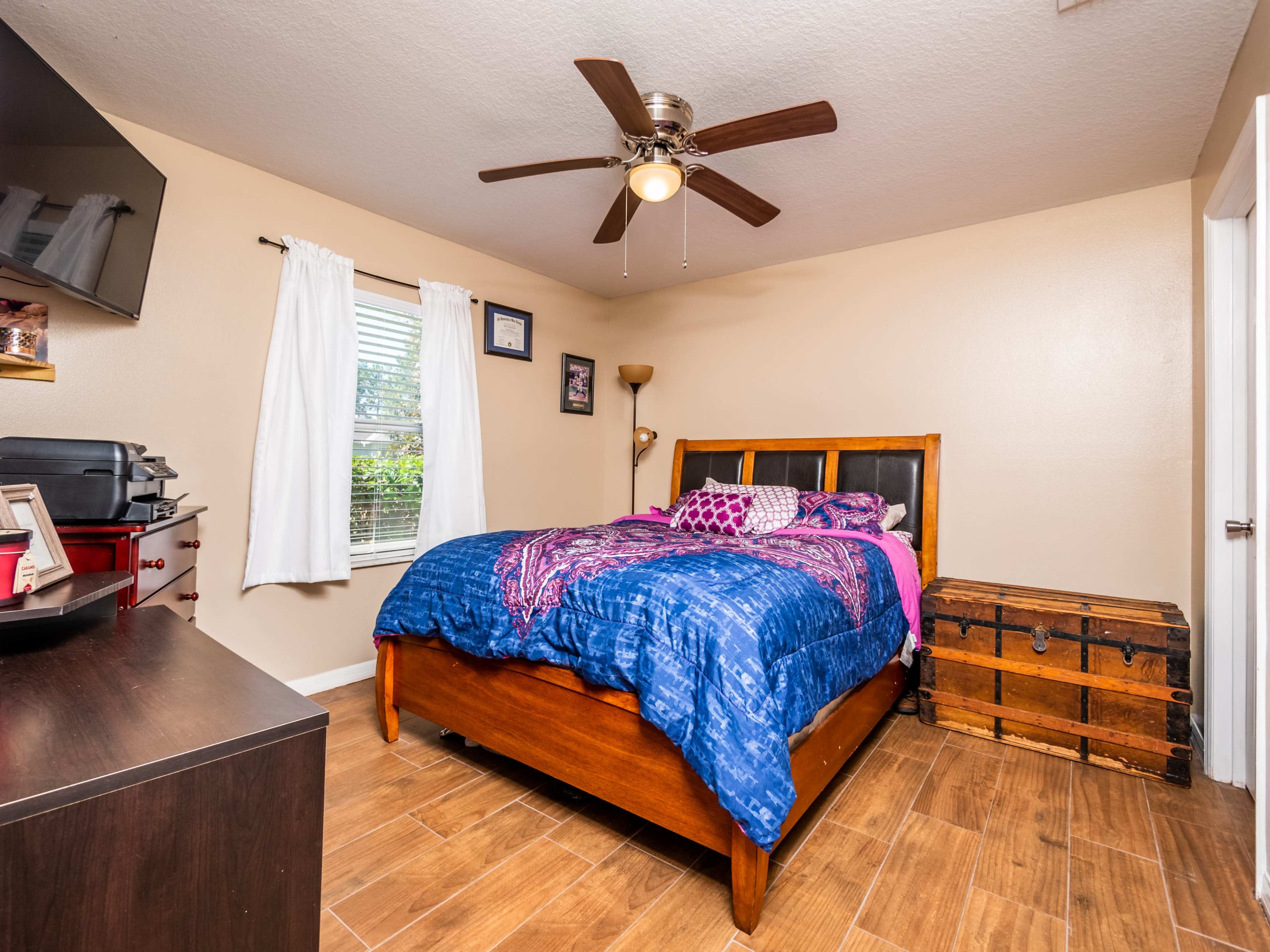 The image shows a bedroom with a wooden bed covered in a blue blanket, a dresser, a wall-mounted television, and a wooden trunk in one corner.