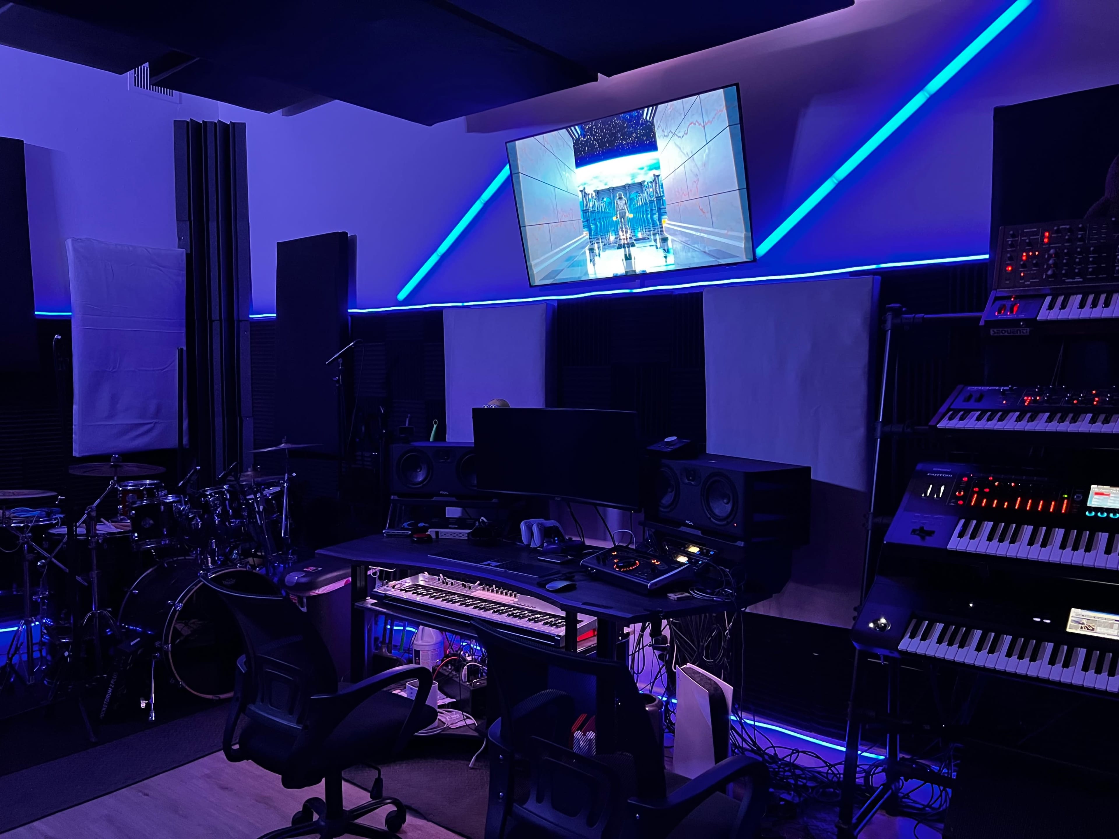 A music studio is equipped with various instruments, a computer setup, and a television displaying a video.