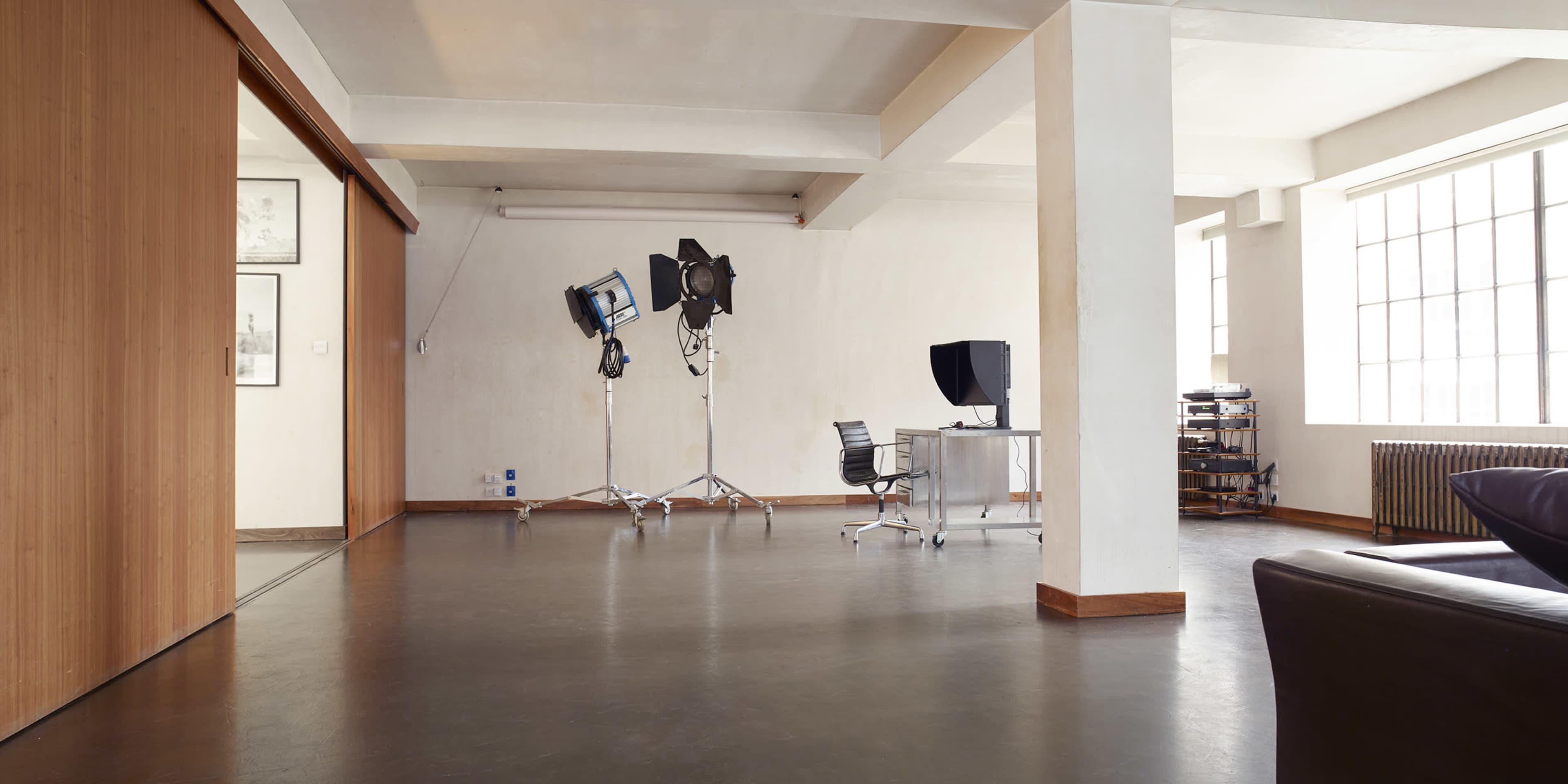 A spacious, minimalistic studio with a desk, a chair, and several lighting equipment positioned in the corner.