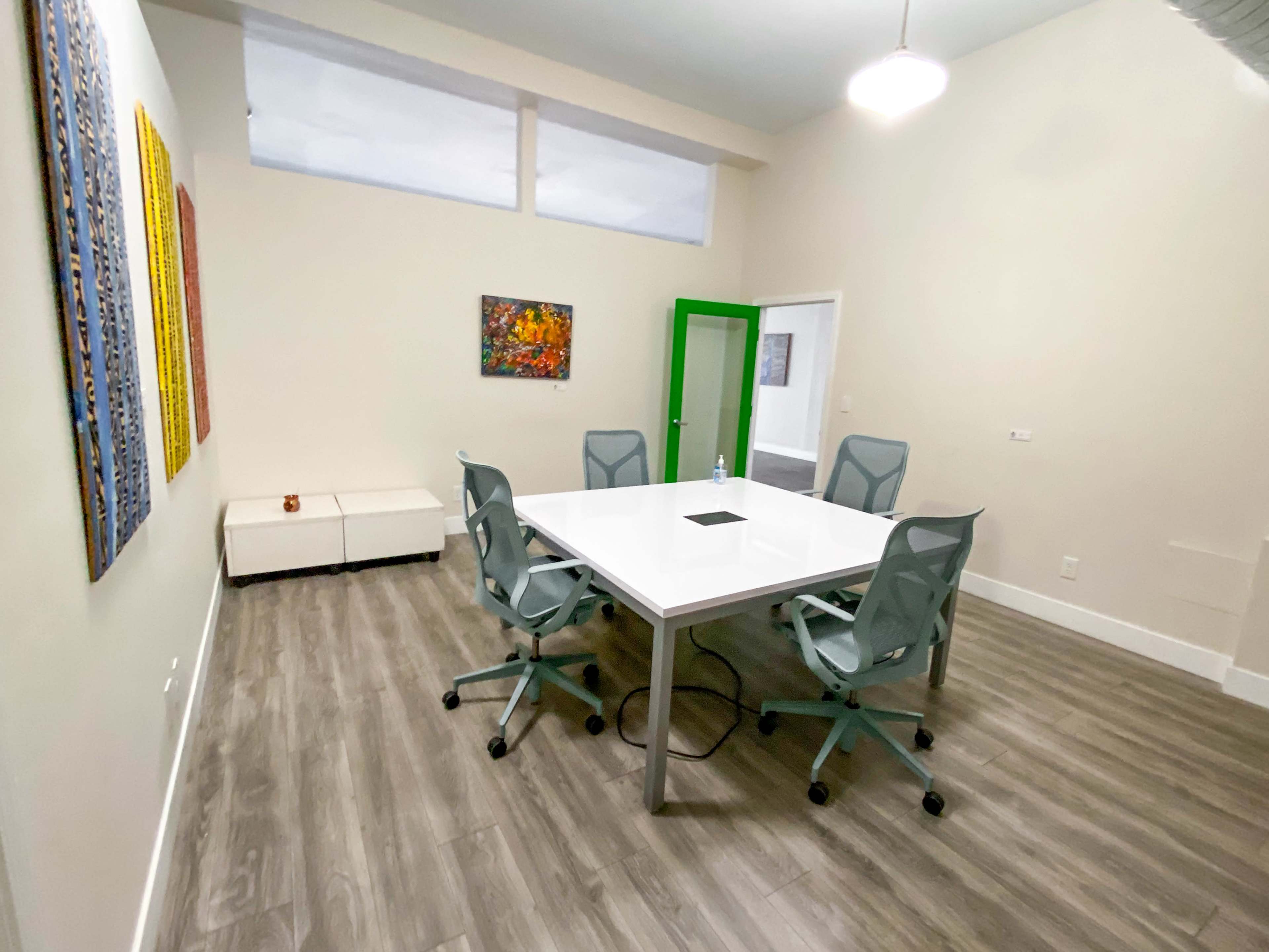 A modern conference room features a white table surrounded by four gray chairs, with abstract artwork on the walls and a green door leading to another space.