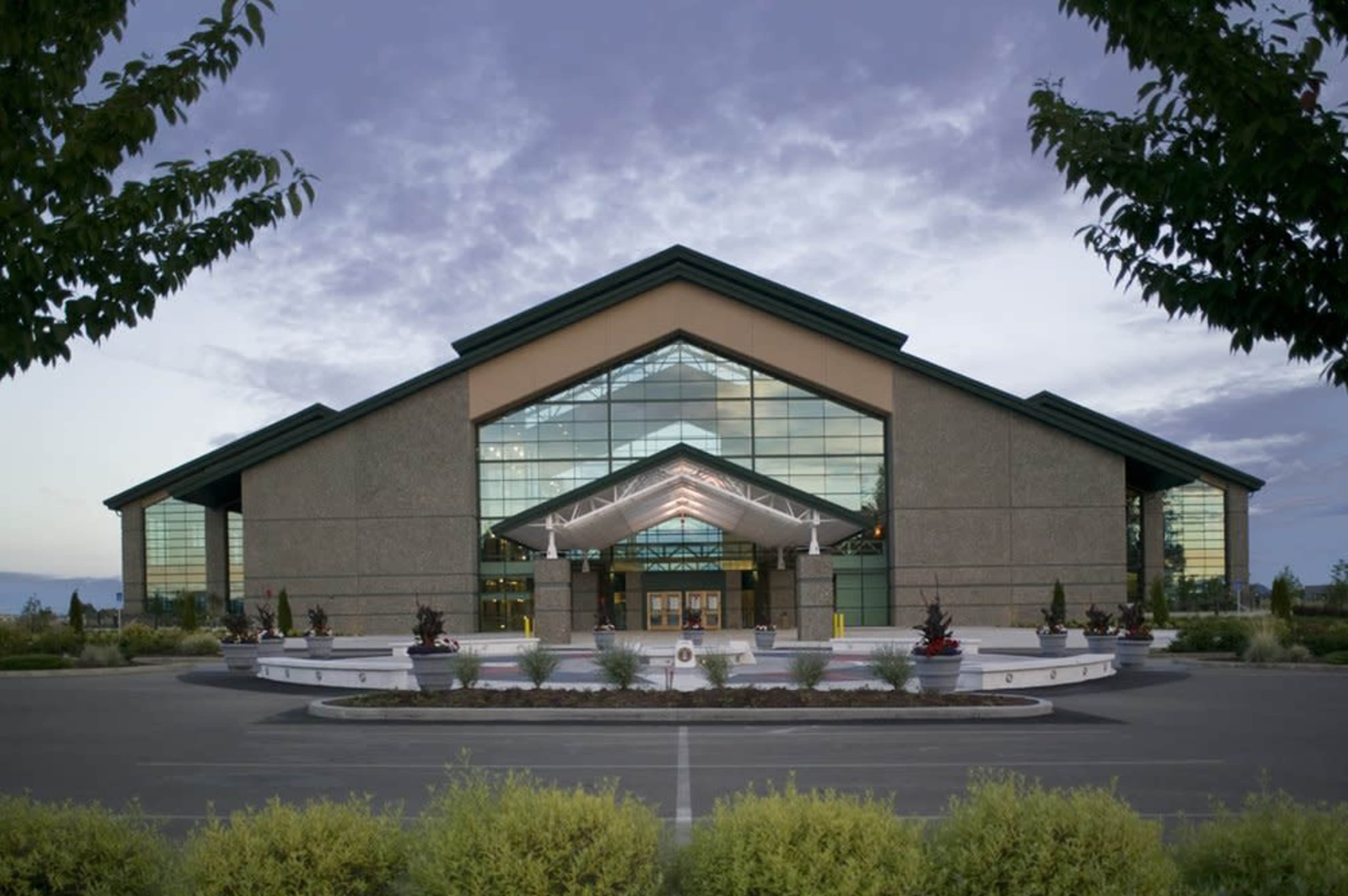 A large, modern building with a peaked roof and extensive glass windows is set against a cloudy sky, surrounded by landscaping and a circular driveway.