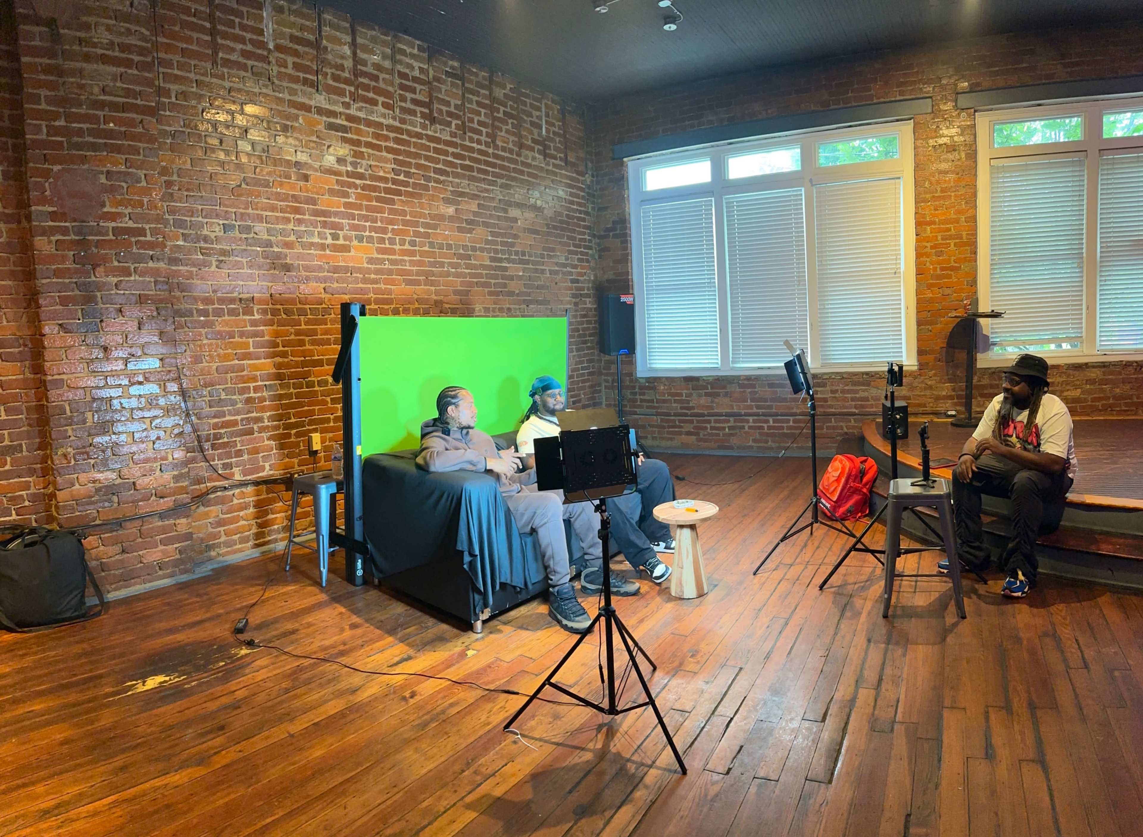 Two individuals sit on a couch in front of a green screen while another person, positioned on the floor, appears to be recording or filming them in a room with exposed brick walls and wooden flooring.