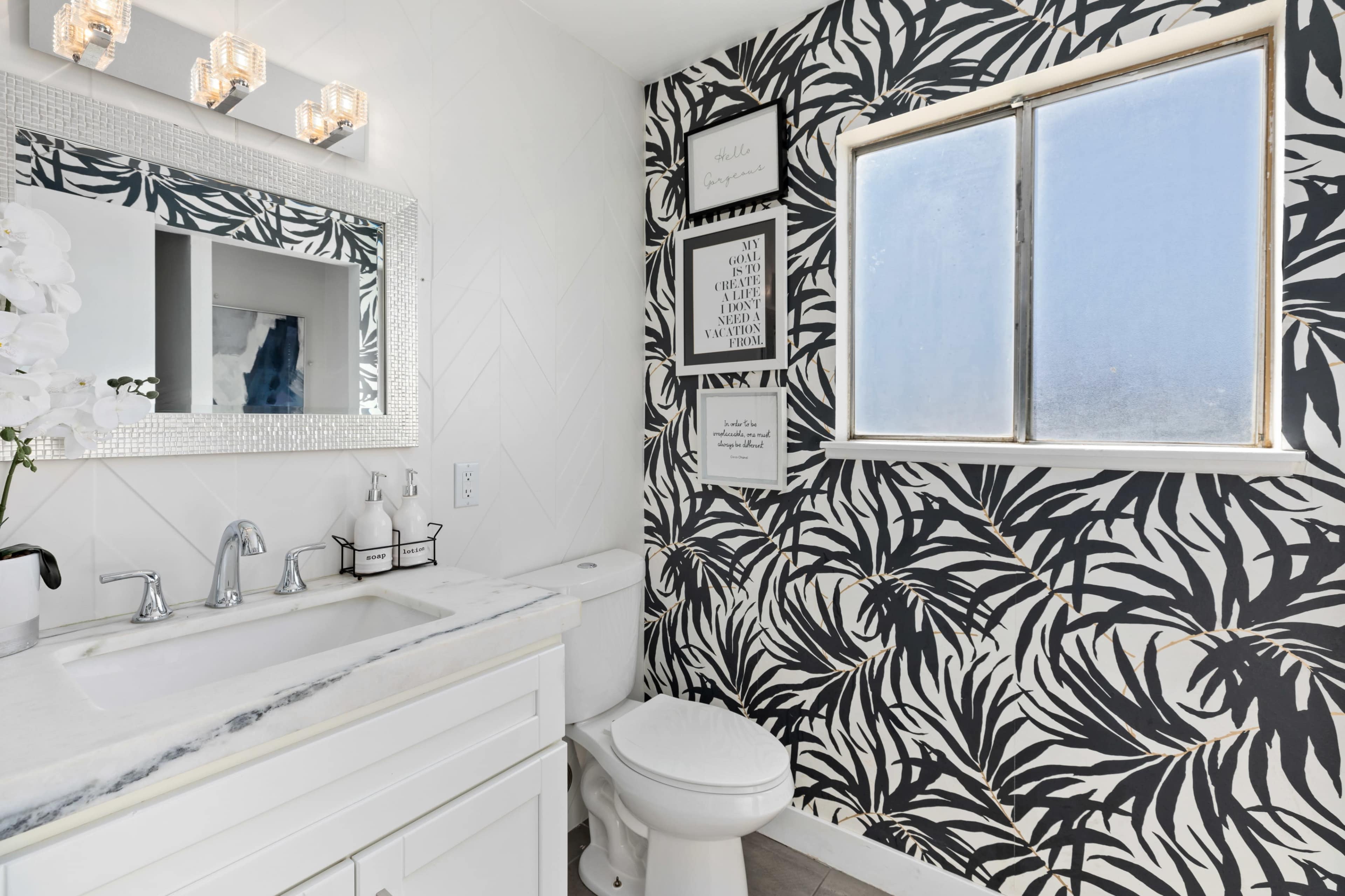 A bathroom with a white vanity, a large mirror, and a black and white tropical leaf-patterned wallpaper.
