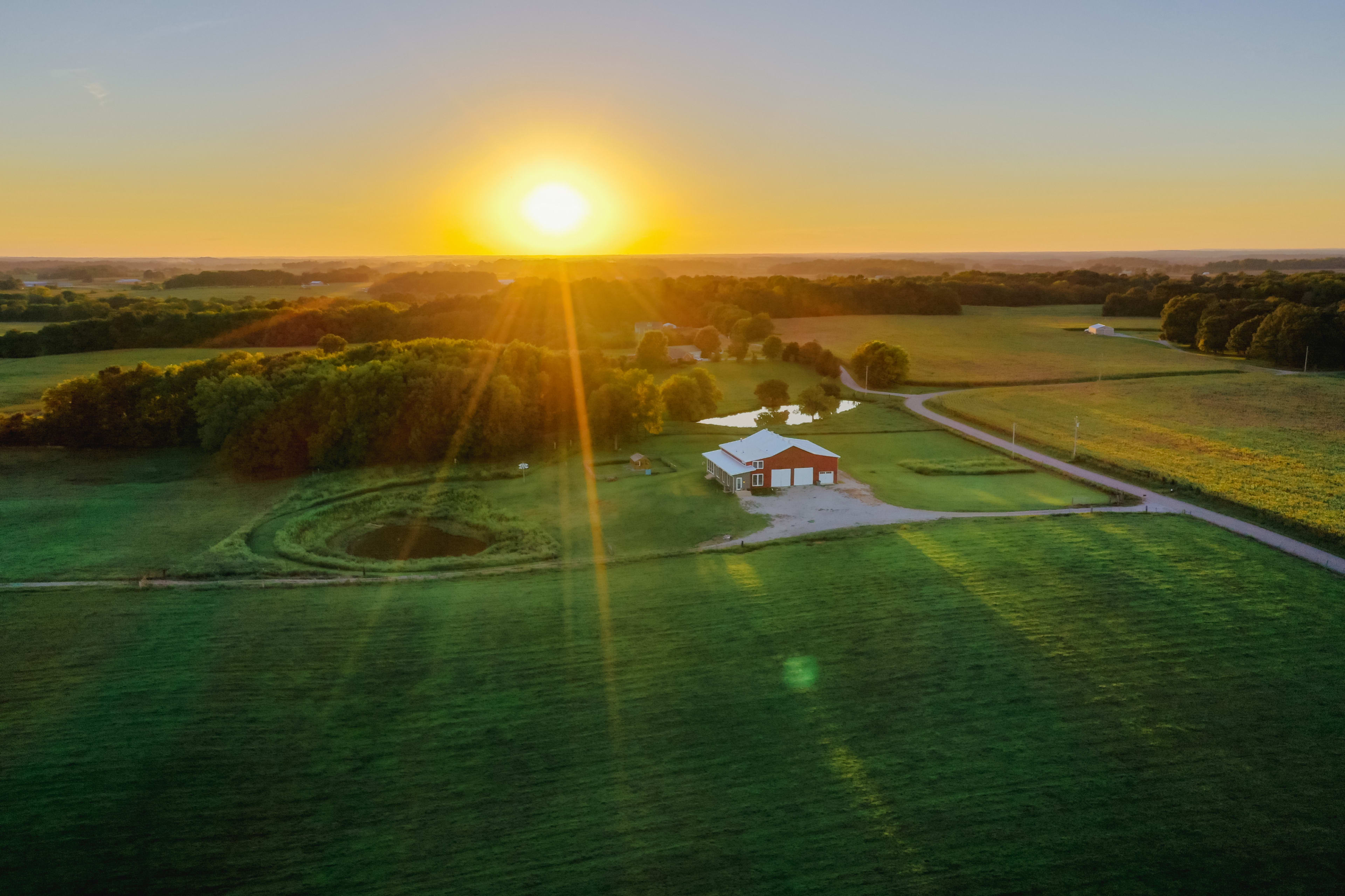 A farmhouse is set in a vast green field with a pond, illuminated by a bright sunrise in the background.