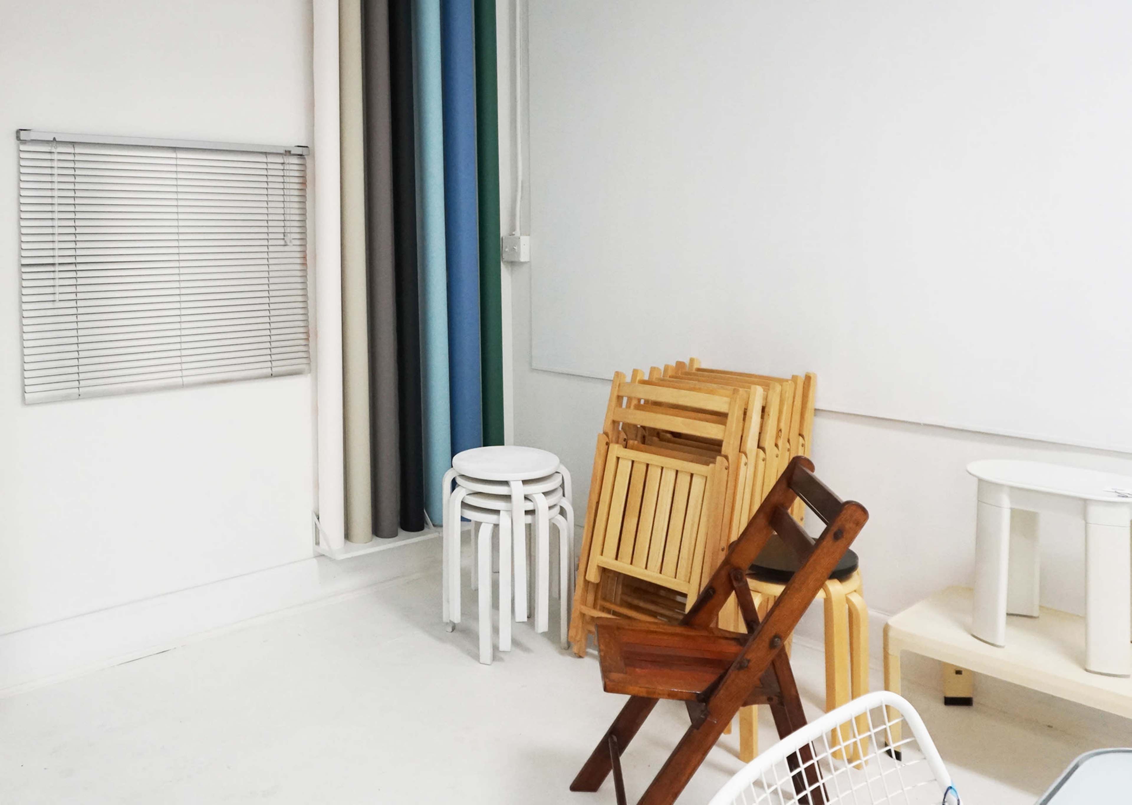 A minimalist room with stacked wooden and folding chairs against a white wall, alongside a window covered with blinds and fabric drapes.