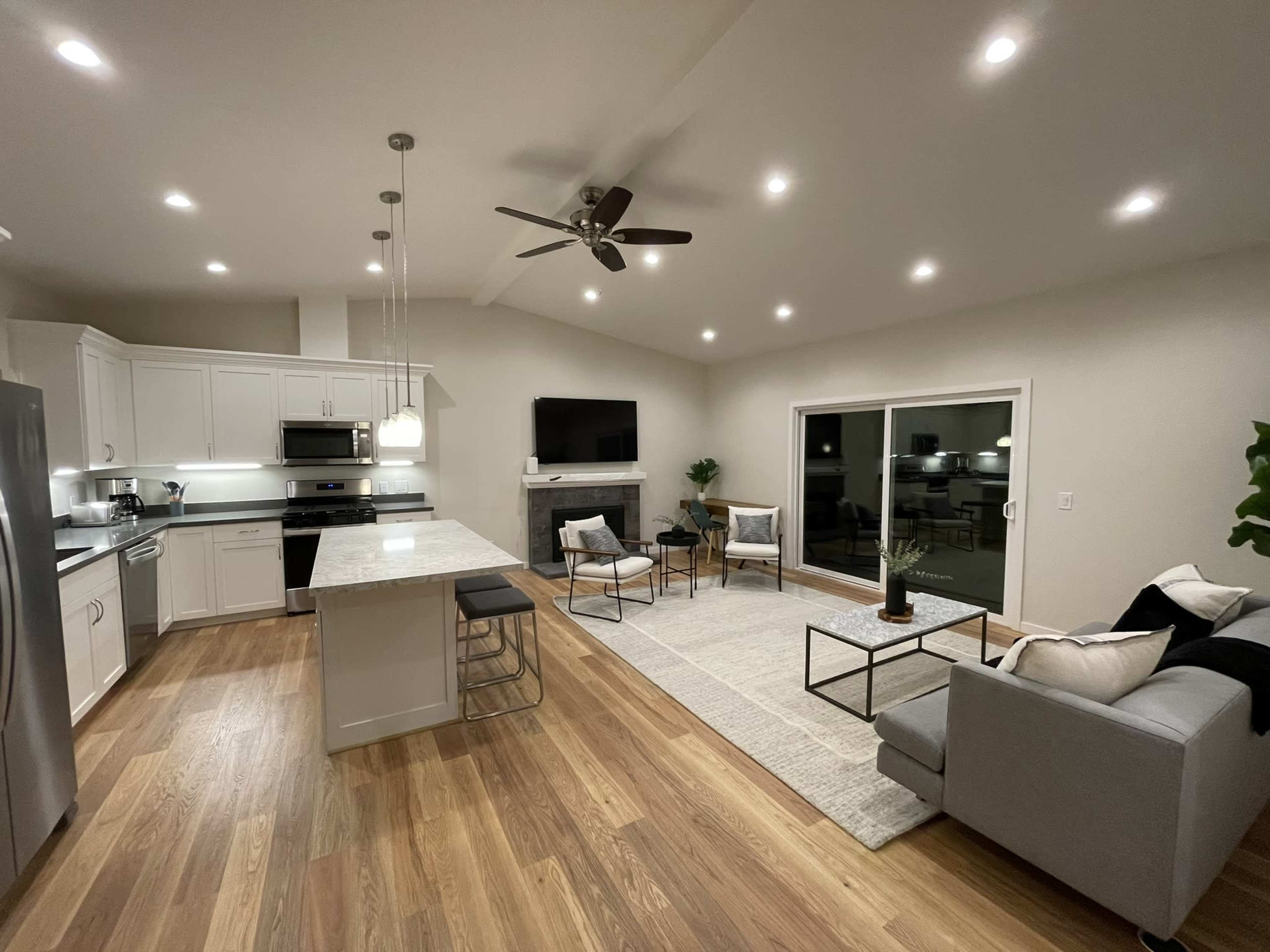 The image shows a modern open-concept living space with a kitchen, dining area, and seating area featuring contemporary furniture and a large window.