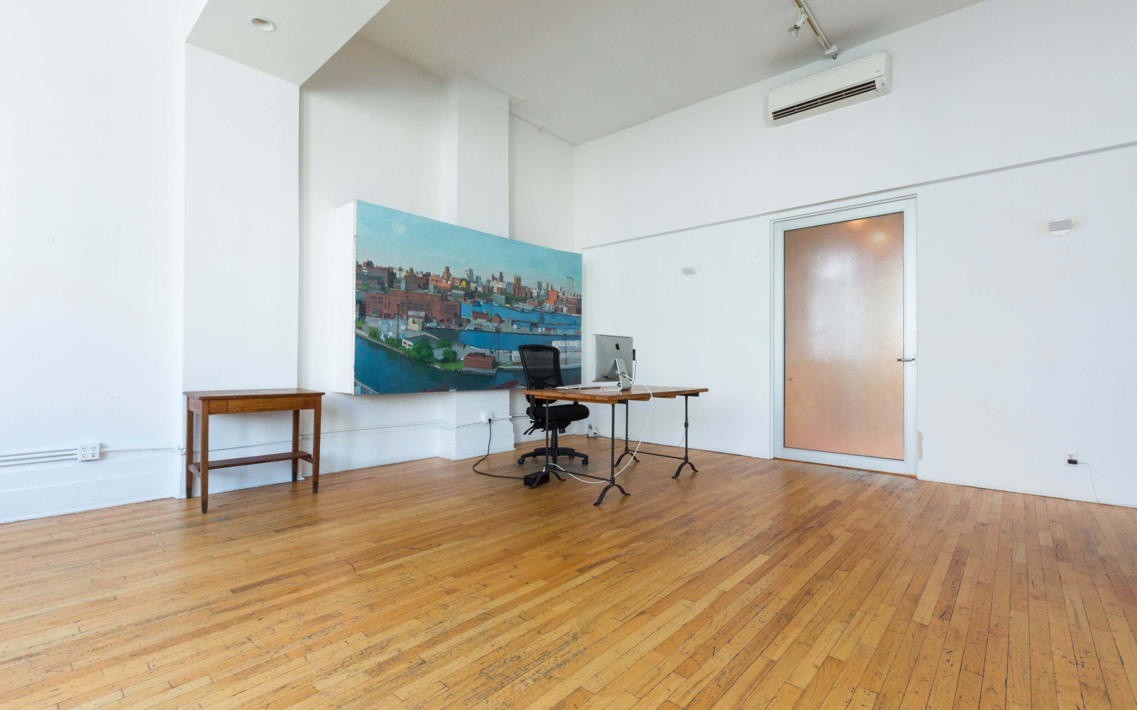 An empty office space features a wooden table with a chair and an artwork on the wall, illuminated by natural light.