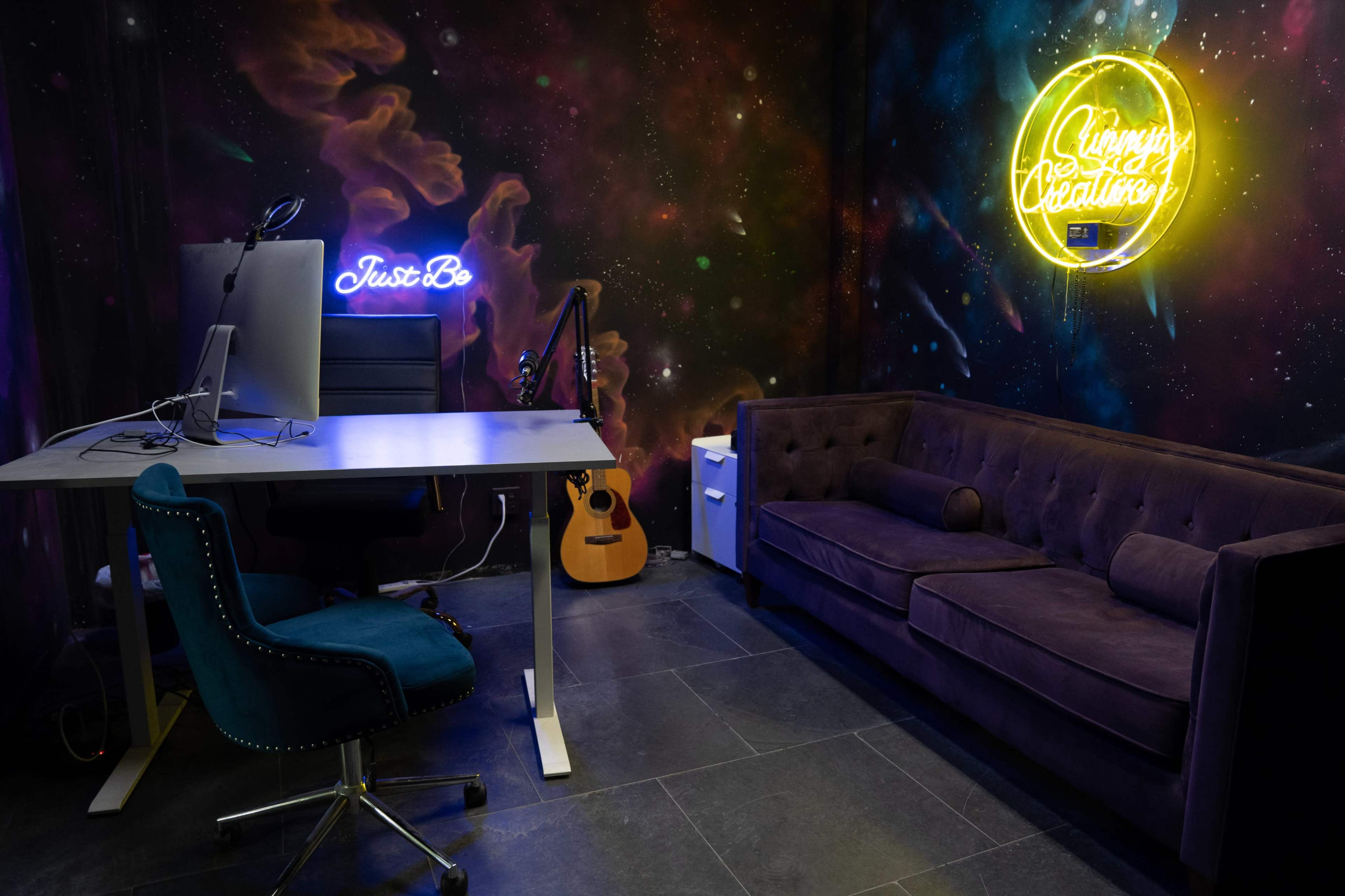 The image shows a modern workspace with a desk, computer, purple couch, and a neon sign on the wall.